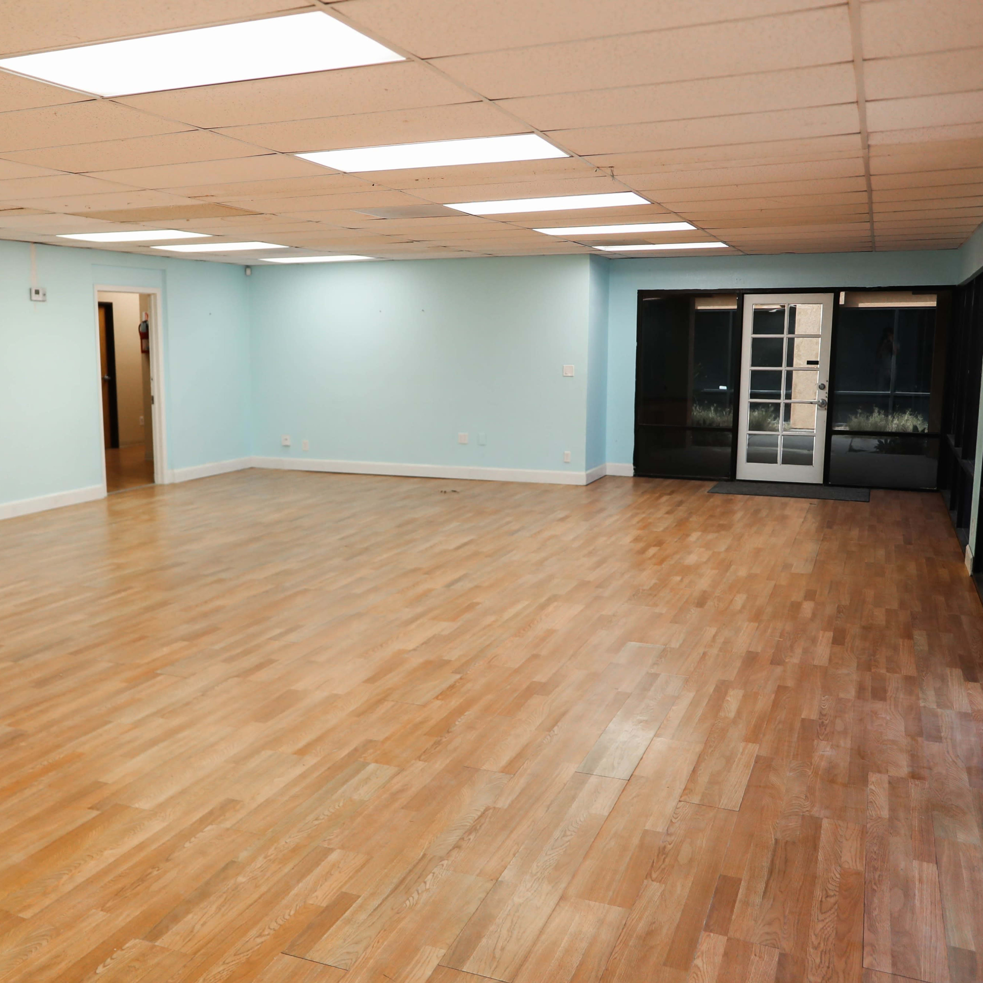 An empty room with light blue walls and hardwood flooring features overhead fluorescent lights and large windows on one side.