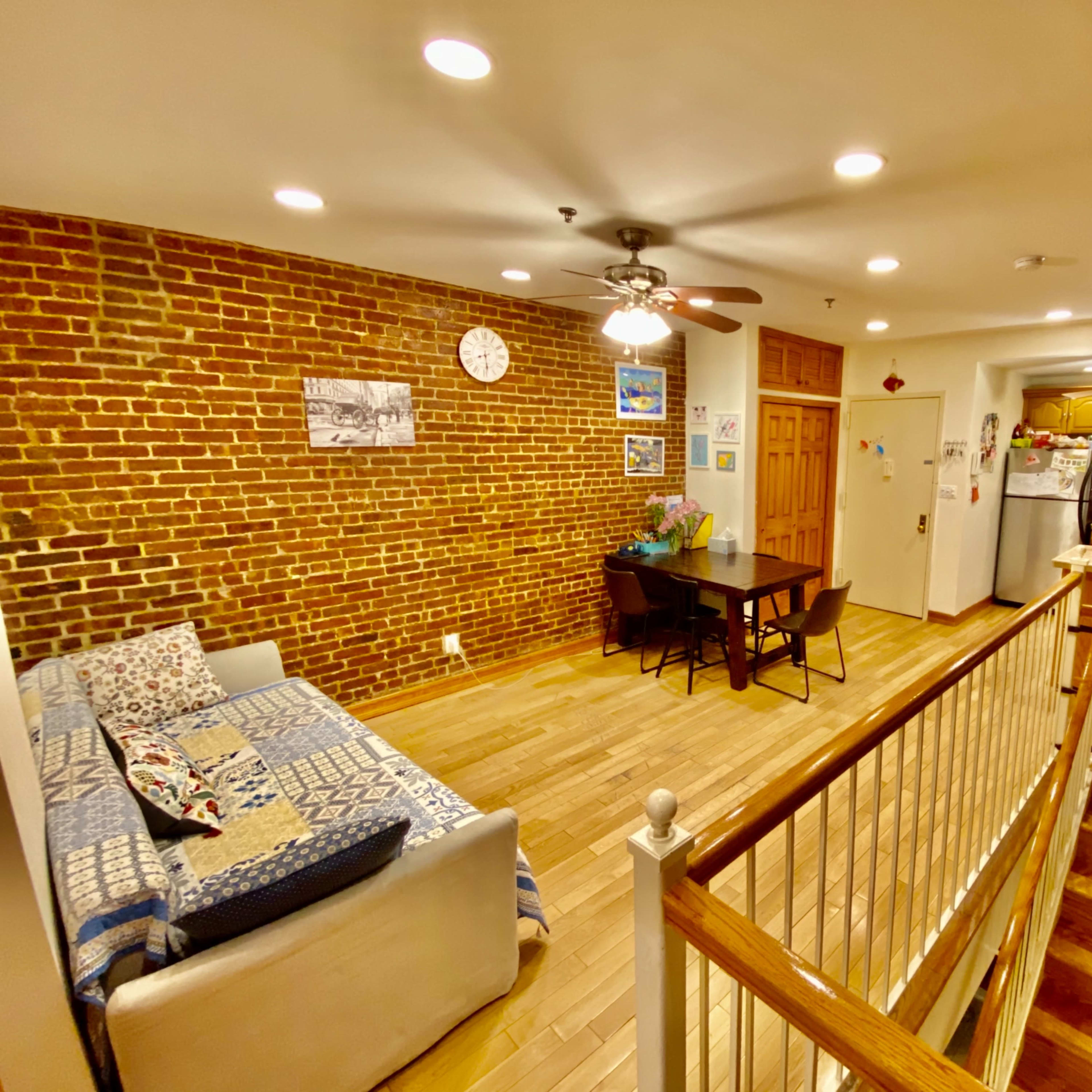 A spacious living area with exposed brick walls, a sofa, a dining table, and a kitchen visible in the background.