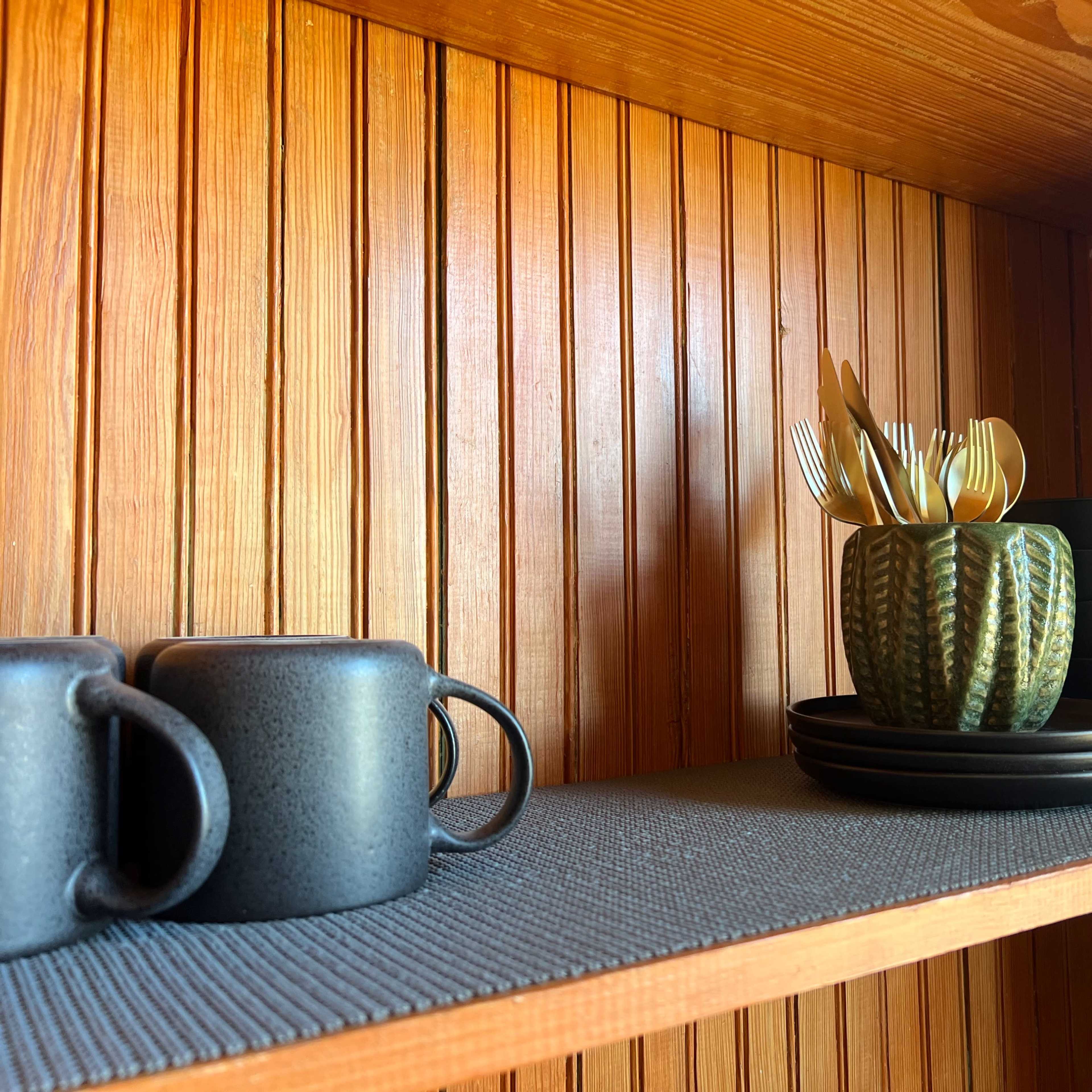 A wooden shelf with black ceramic mugs and plates, a green cactus-shaped container holding golden utensils, and a textured runner.