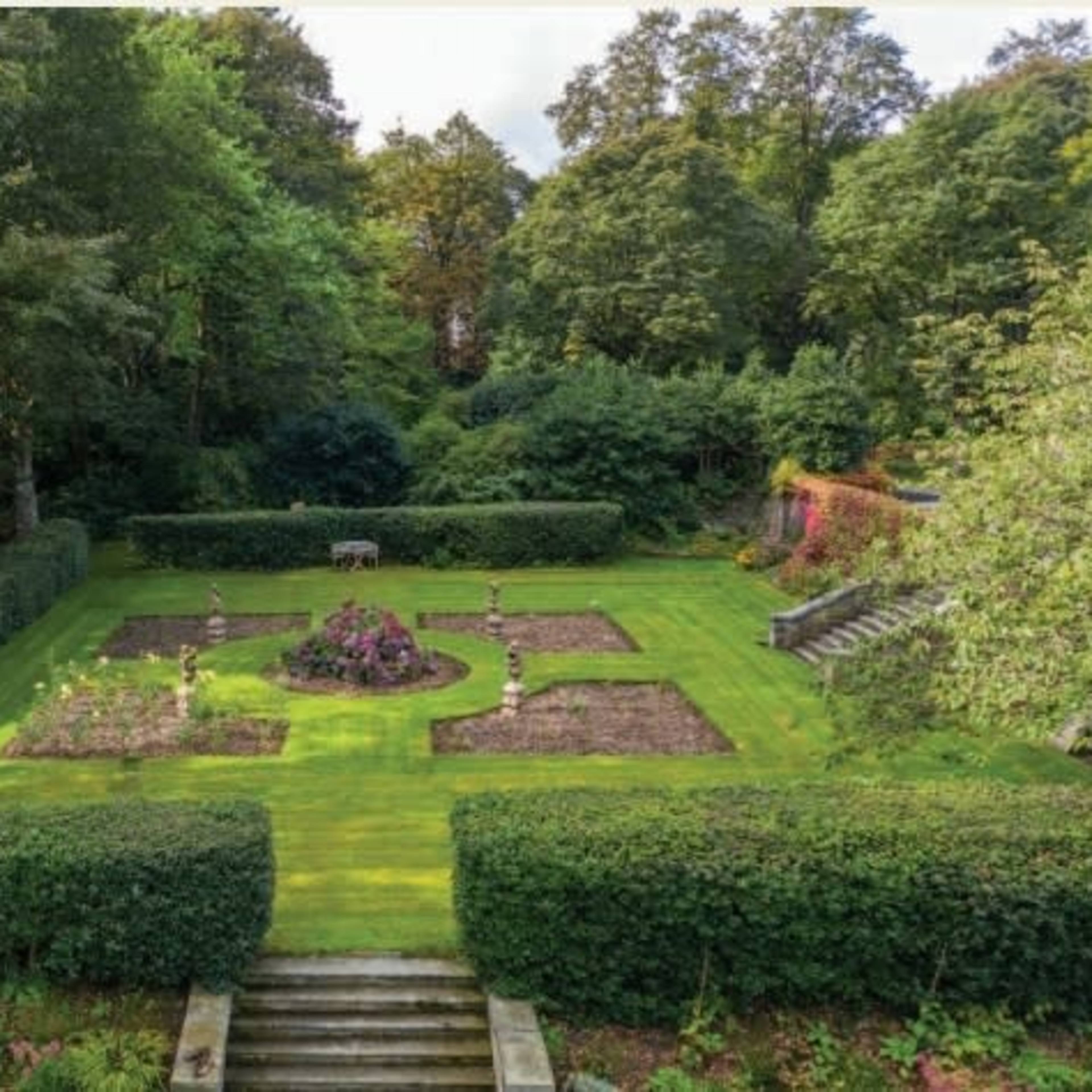 A well-maintained garden surrounded by neatly trimmed hedges, featuring flowerbeds arranged in a circular pattern and a set of stone steps leading into the garden.