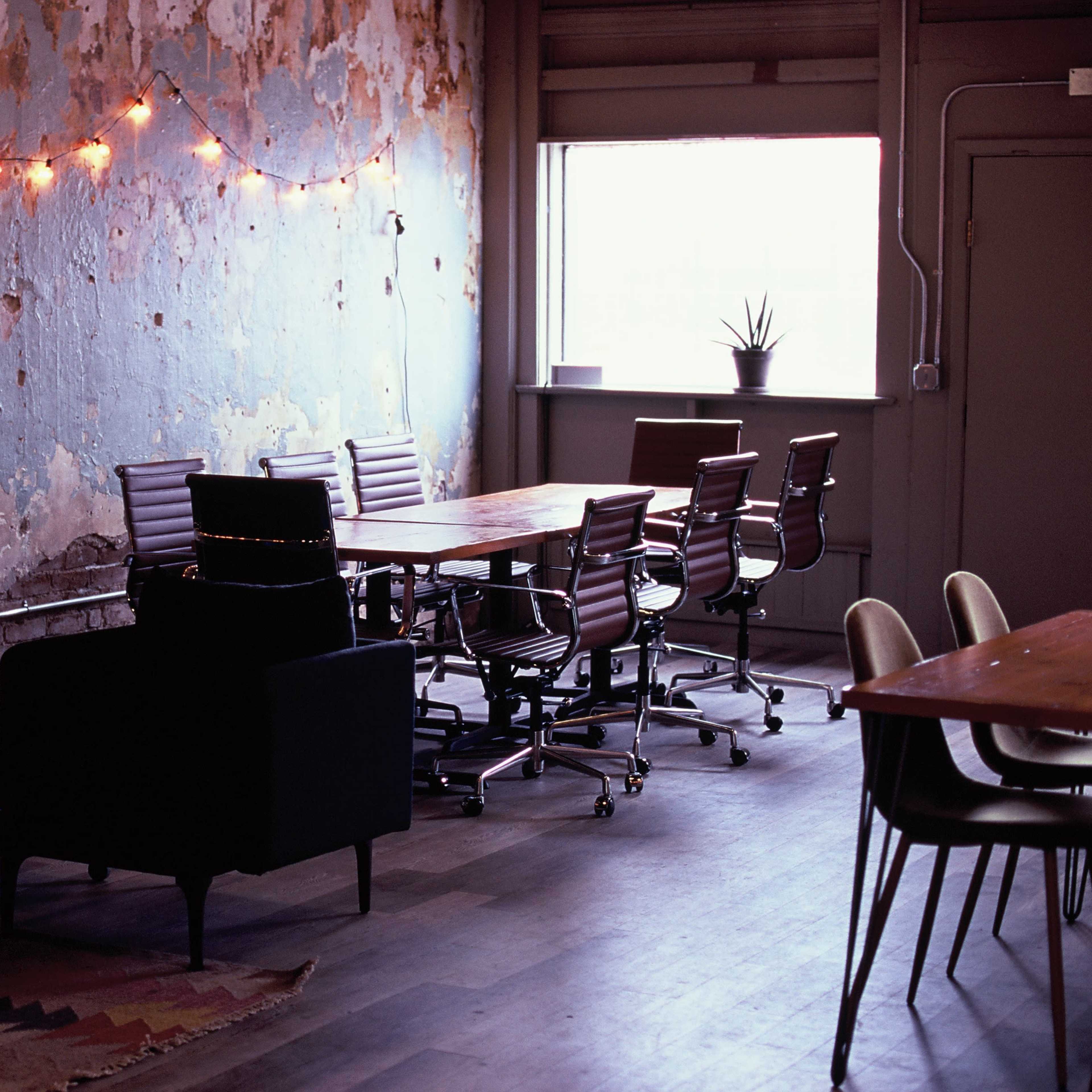 A workspace features a mix of chairs and tables against a weathered blue wall illuminated by string lights.