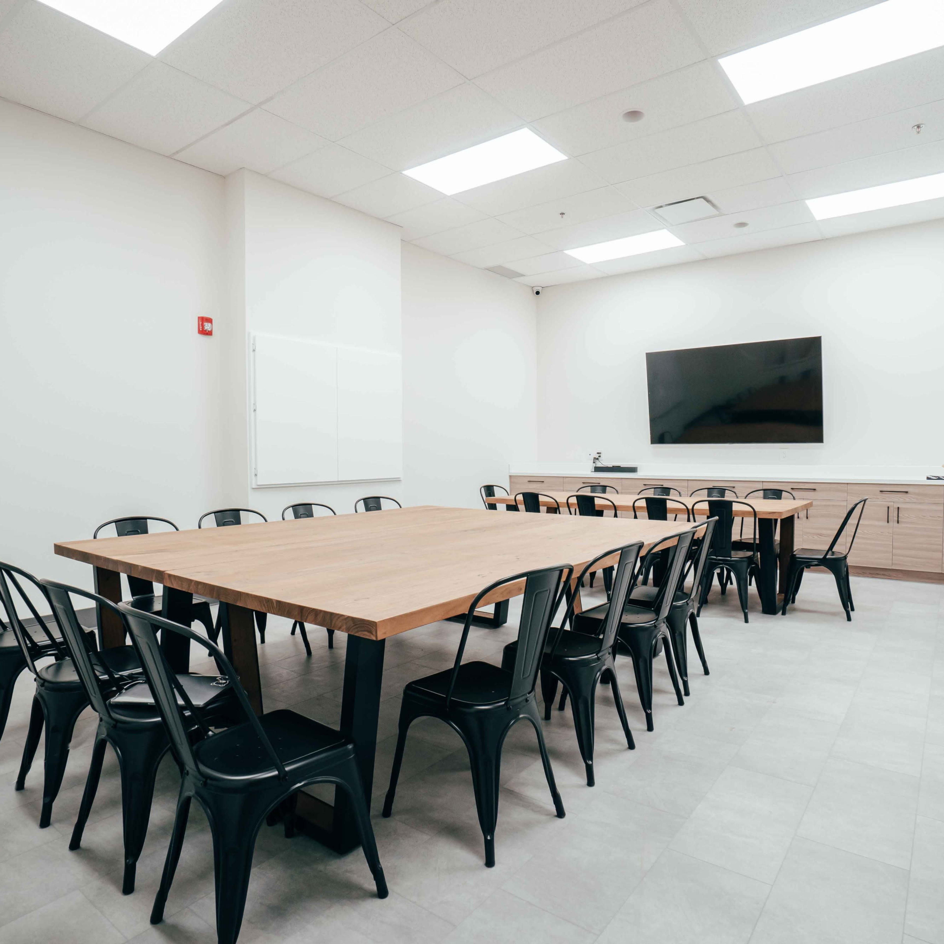 A vacant meeting room features a large wooden table surrounded by black chairs, with a wall-mounted screen and minimal furnishings.