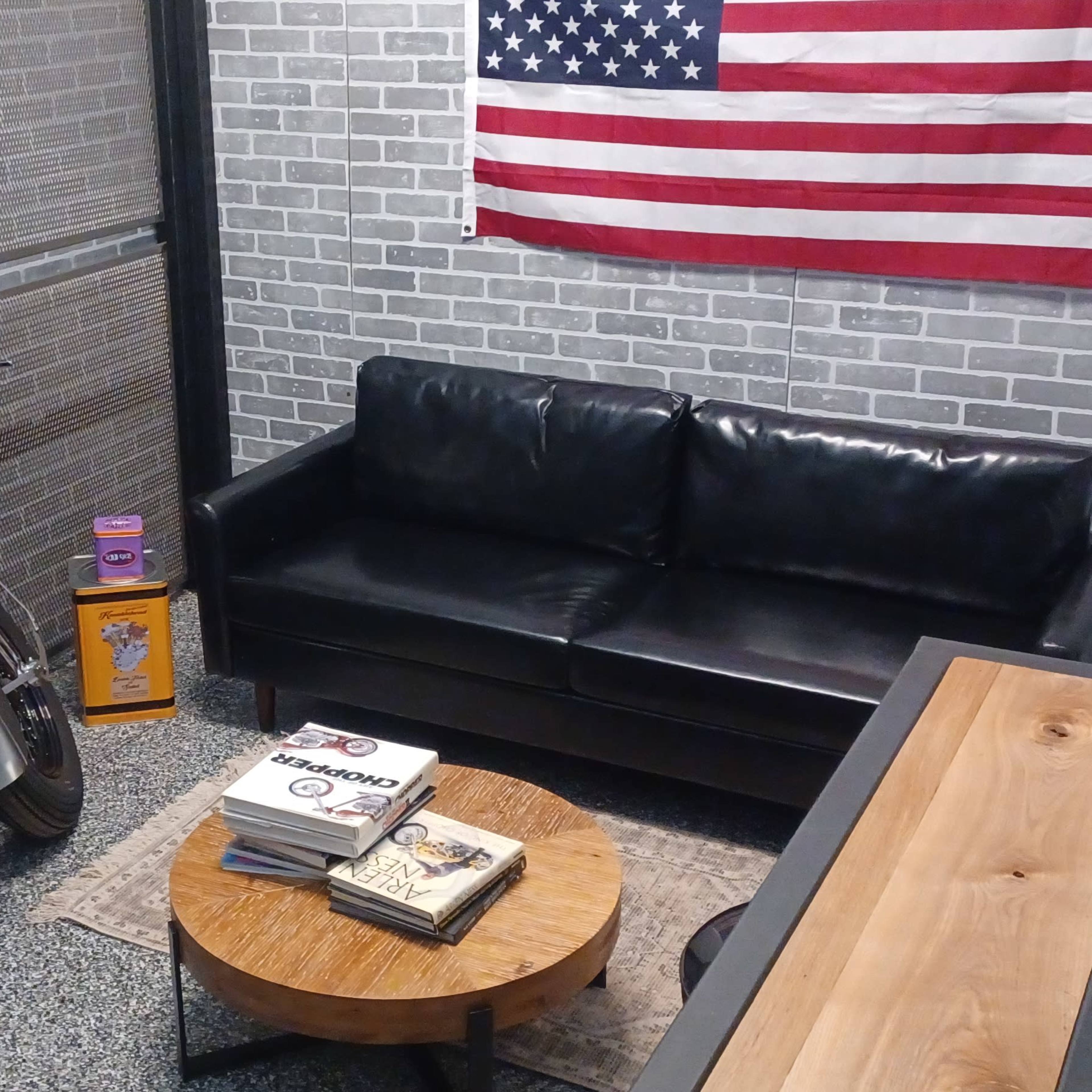 A modern room with a motorcycle parked beside a black leather sofa, a wooden table in the center, and an American flag hanging on the wall.