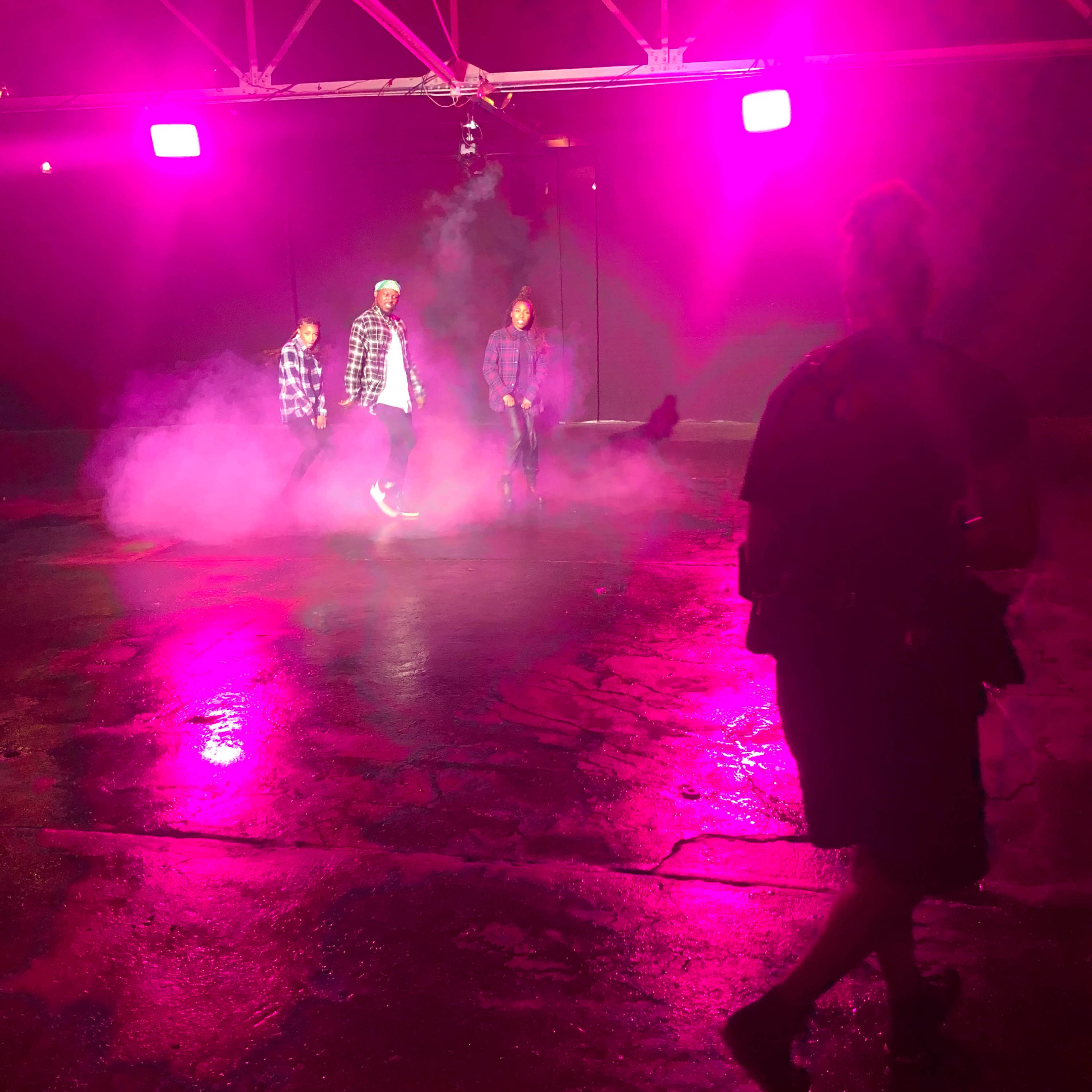 Three individuals stand on a reflective surface surrounded by pink lighting and fog.
