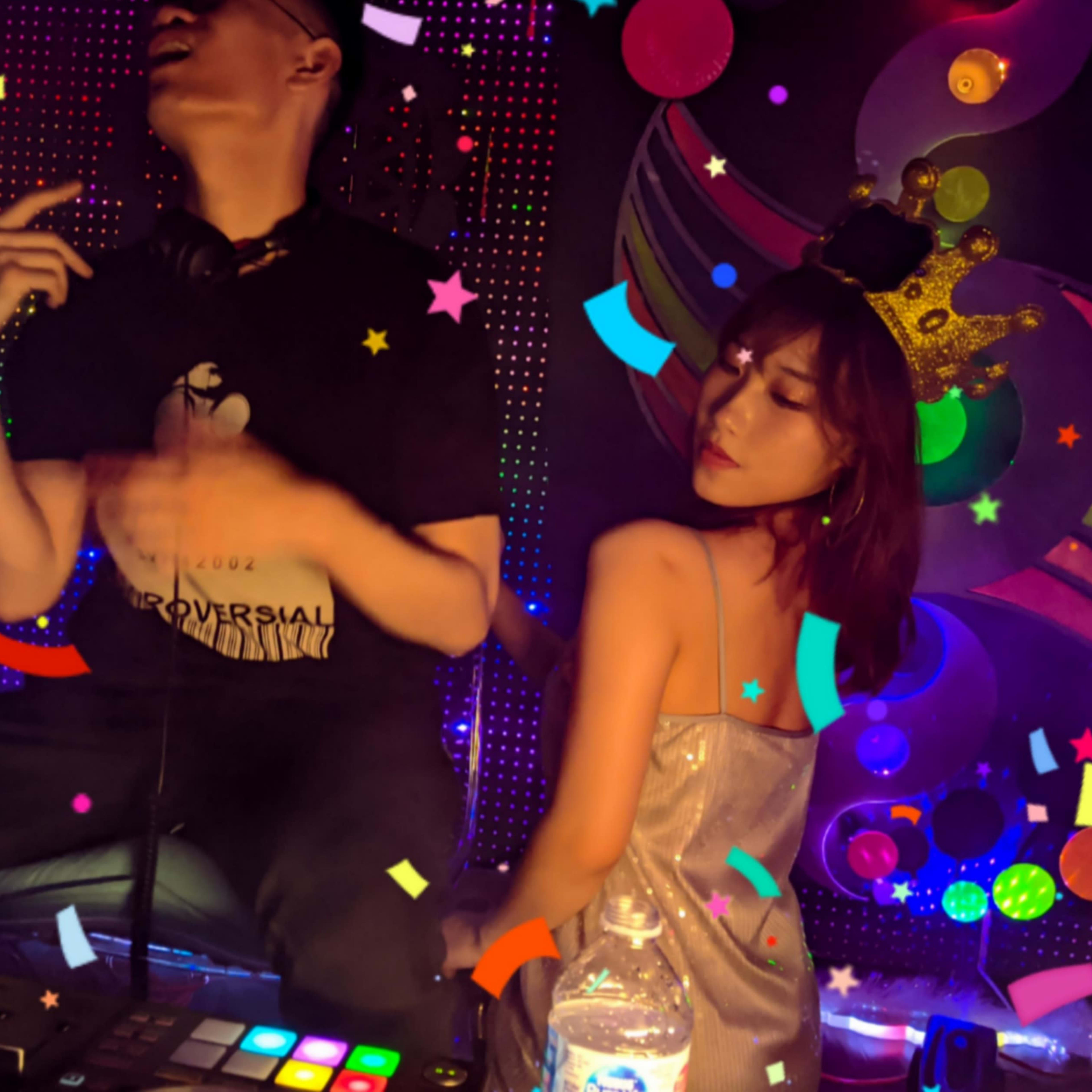 A DJ performs at a lively event while a woman in a shimmering dress poses next to him, surrounded by colorful confetti and decorations.