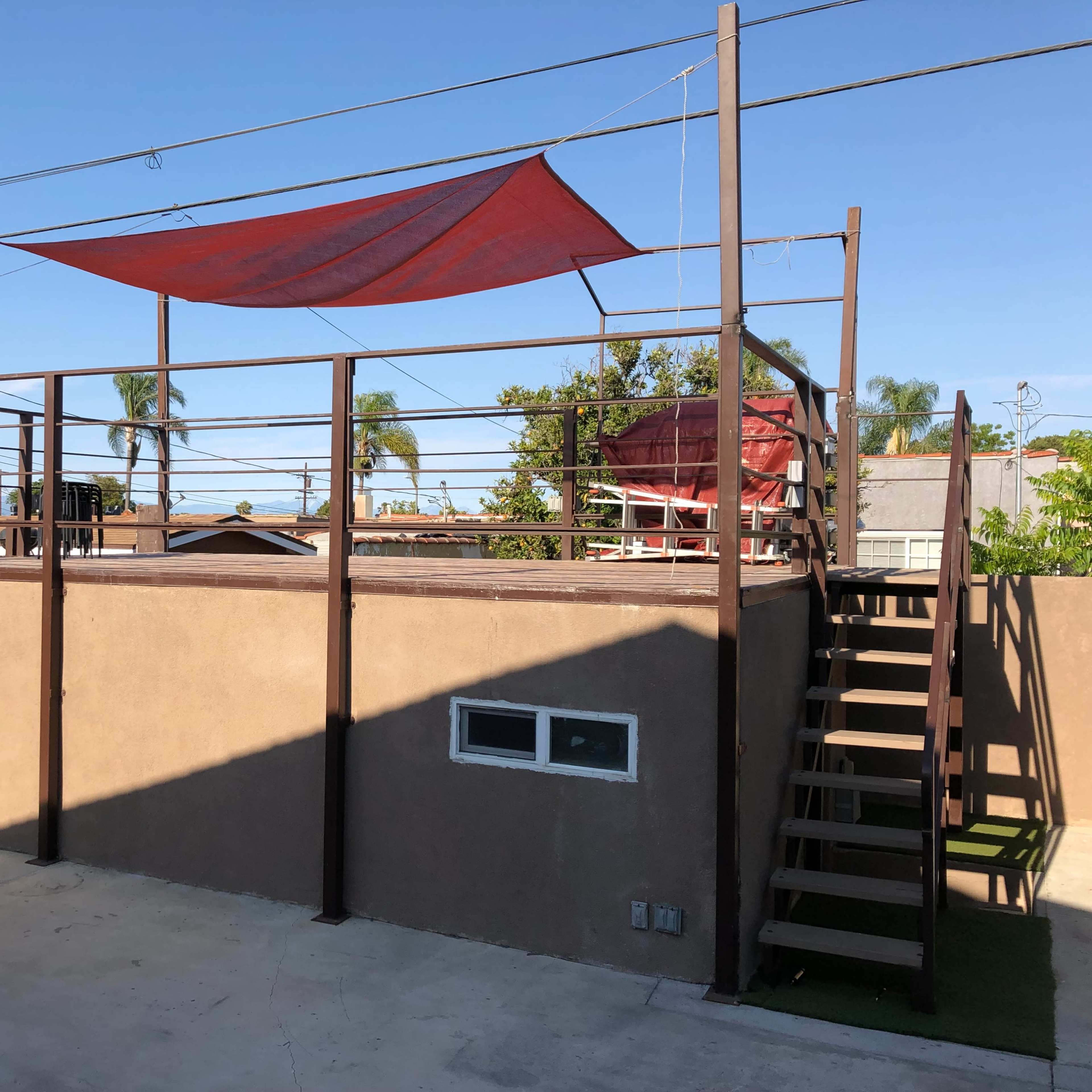 A rooftop area with a red shade sail, surrounded by metal railings, and accessed by a staircase on one side.
