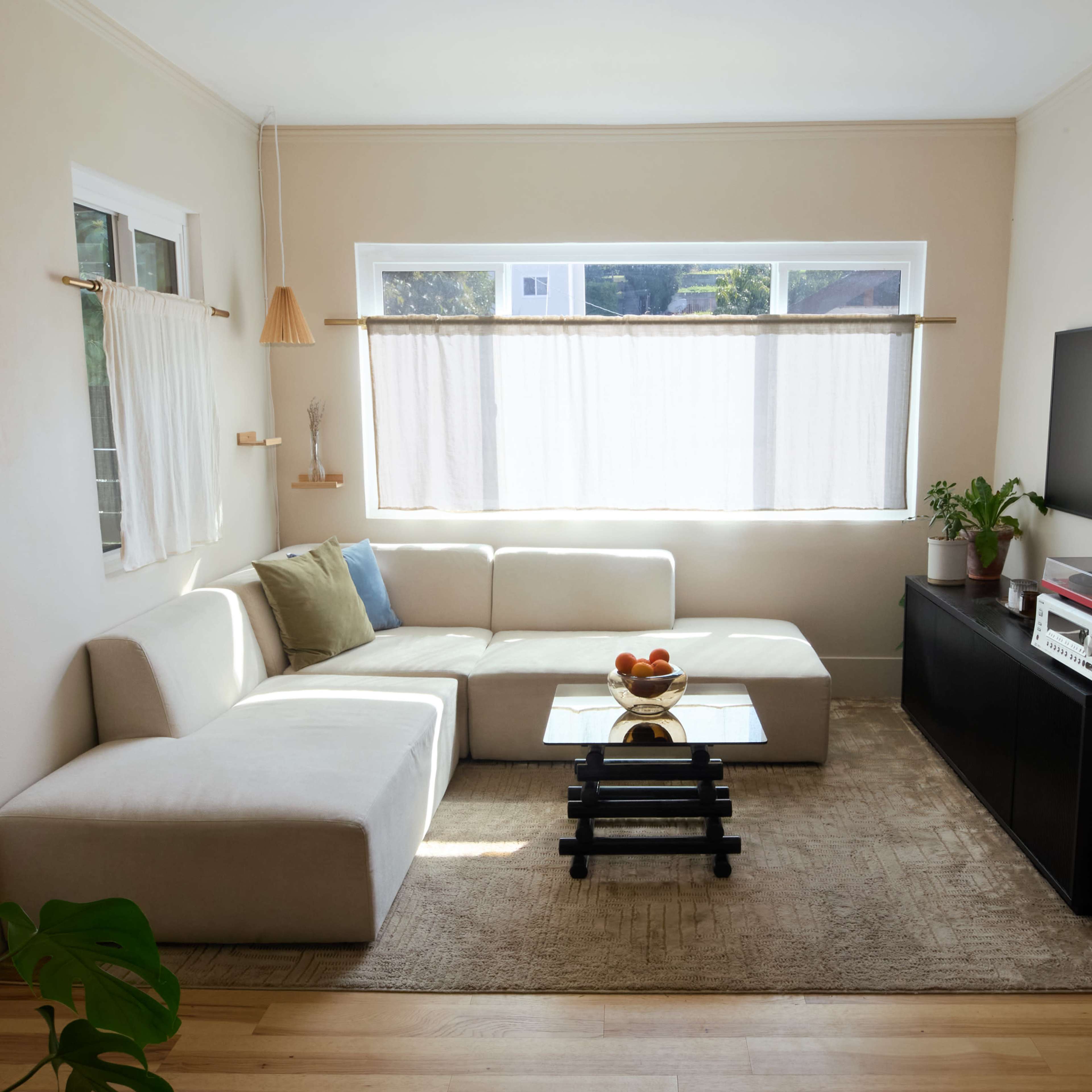 A modern living room featuring a sectional sofa, a glass coffee table, a TV stand with electronic equipment, and natural light filtering through sheer curtains.