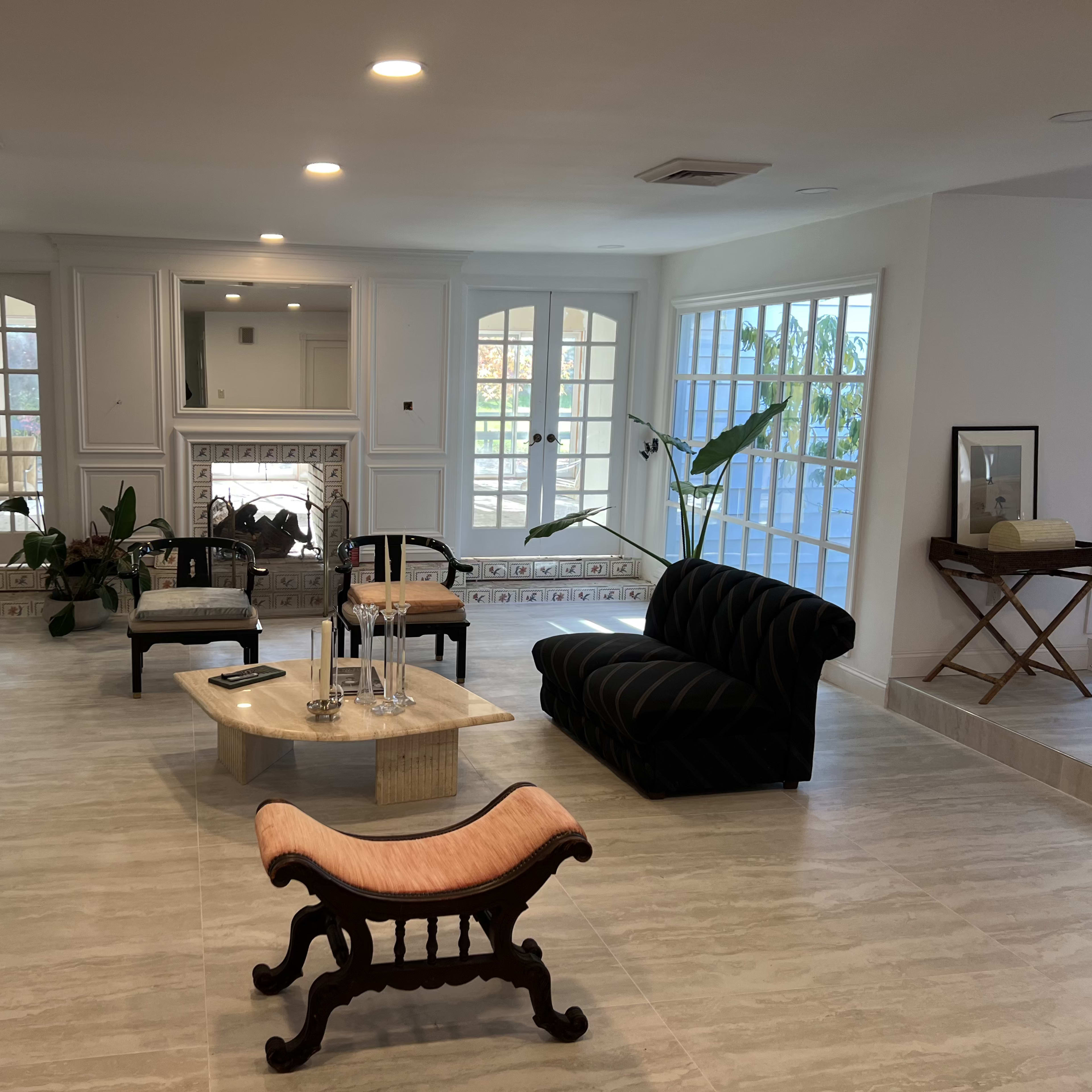 A spacious living room with large windows, a marble coffee table, a black couch, and a wooden bench, surrounded by light-colored tile flooring.