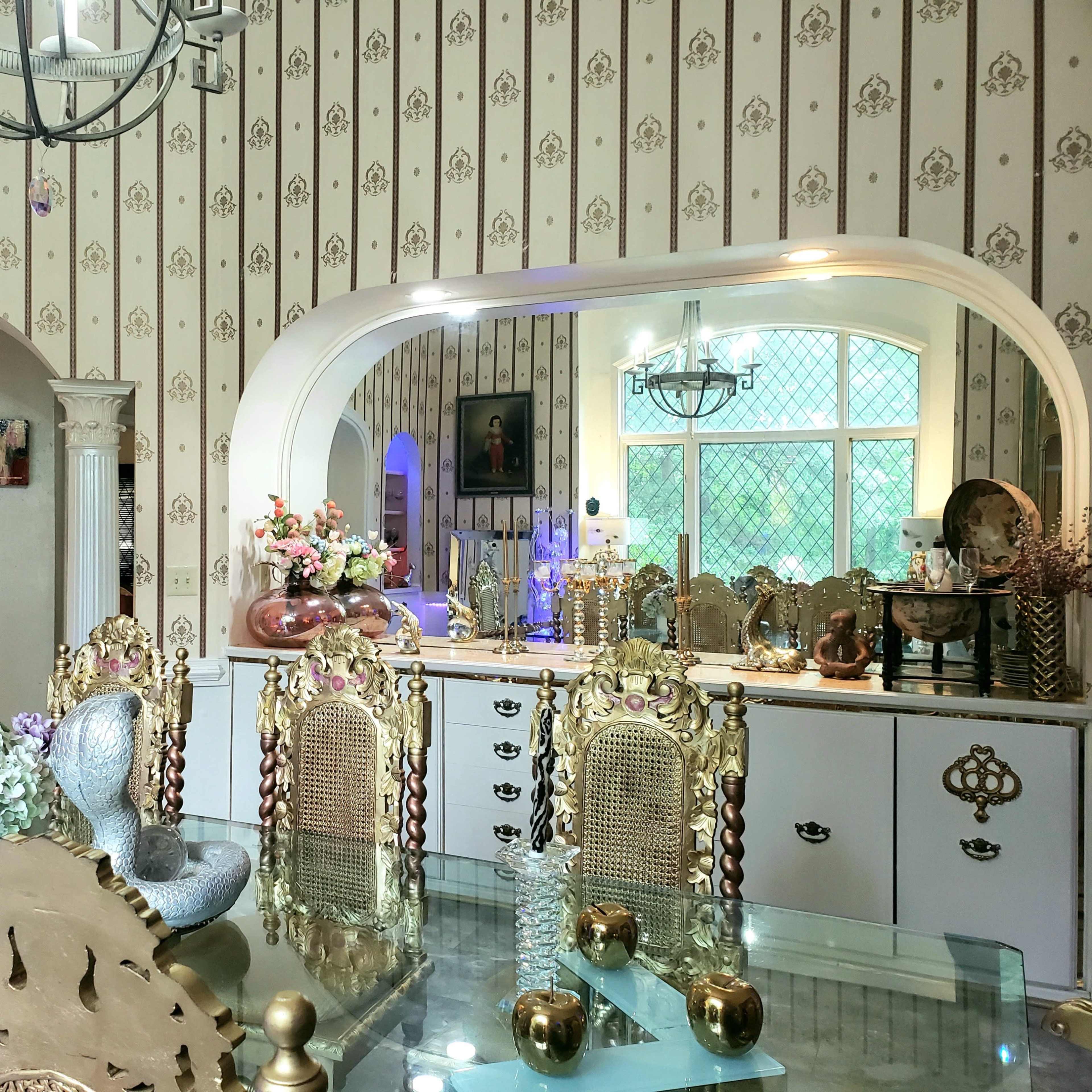 A dining area with ornate chairs, a glass table, and a kitchen visible through an archway, all set against patterned wallpaper.
