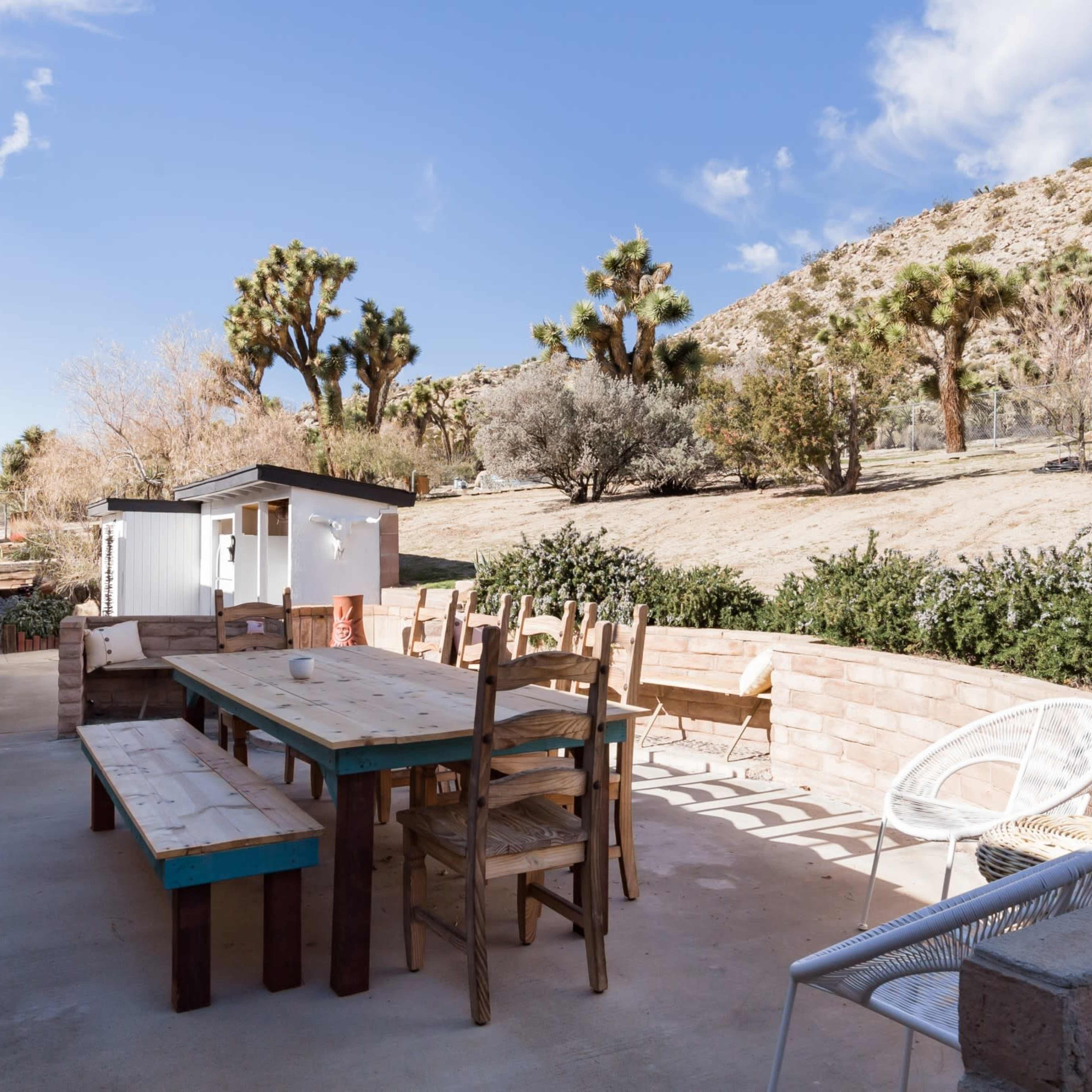 A large outdoor patio features a wooden dining table surrounded by chairs, with desert scenery and shrubs in the background.
