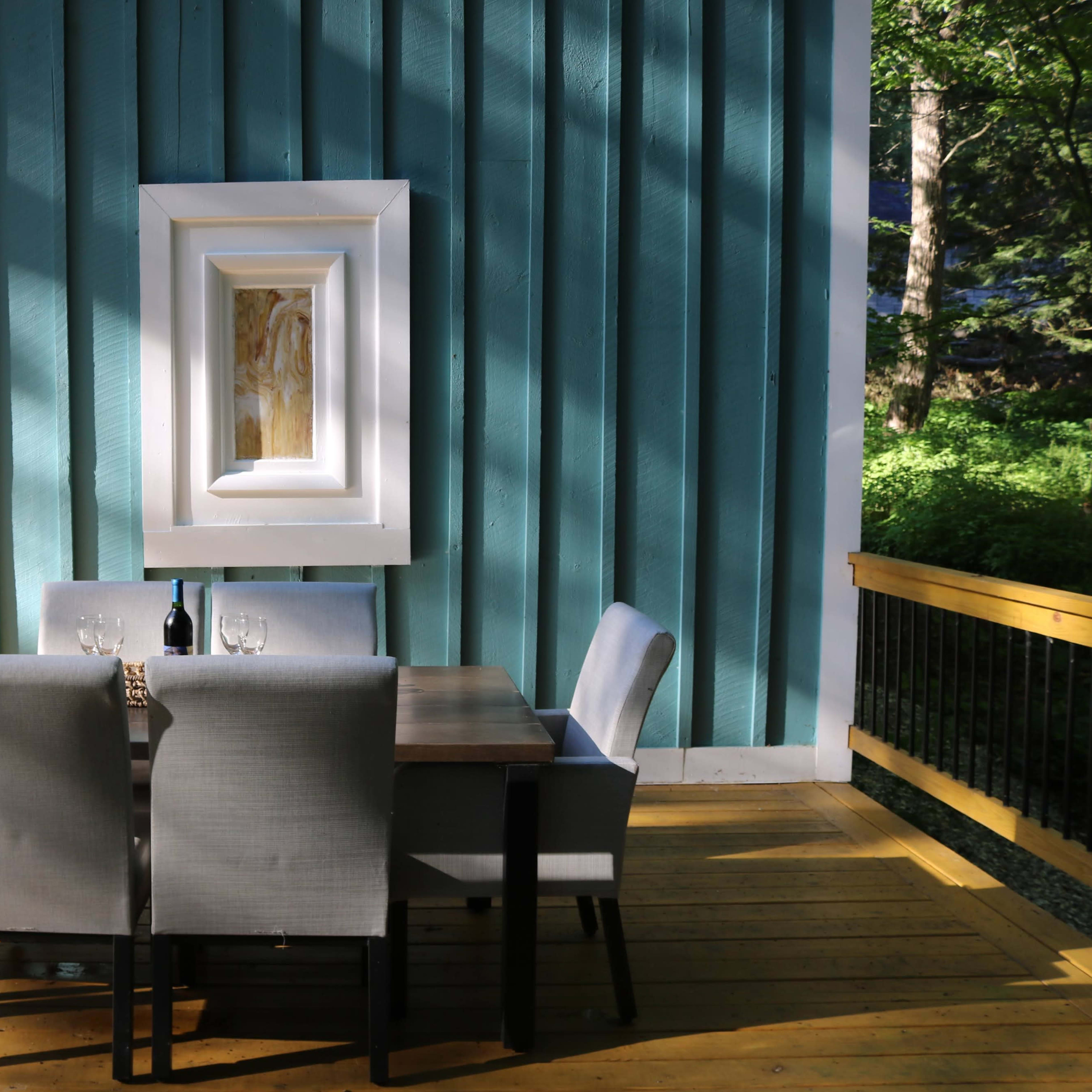 A dining table with gray chairs is situated on a wooden deck against a turquoise wall, featuring a framed painting above the table.