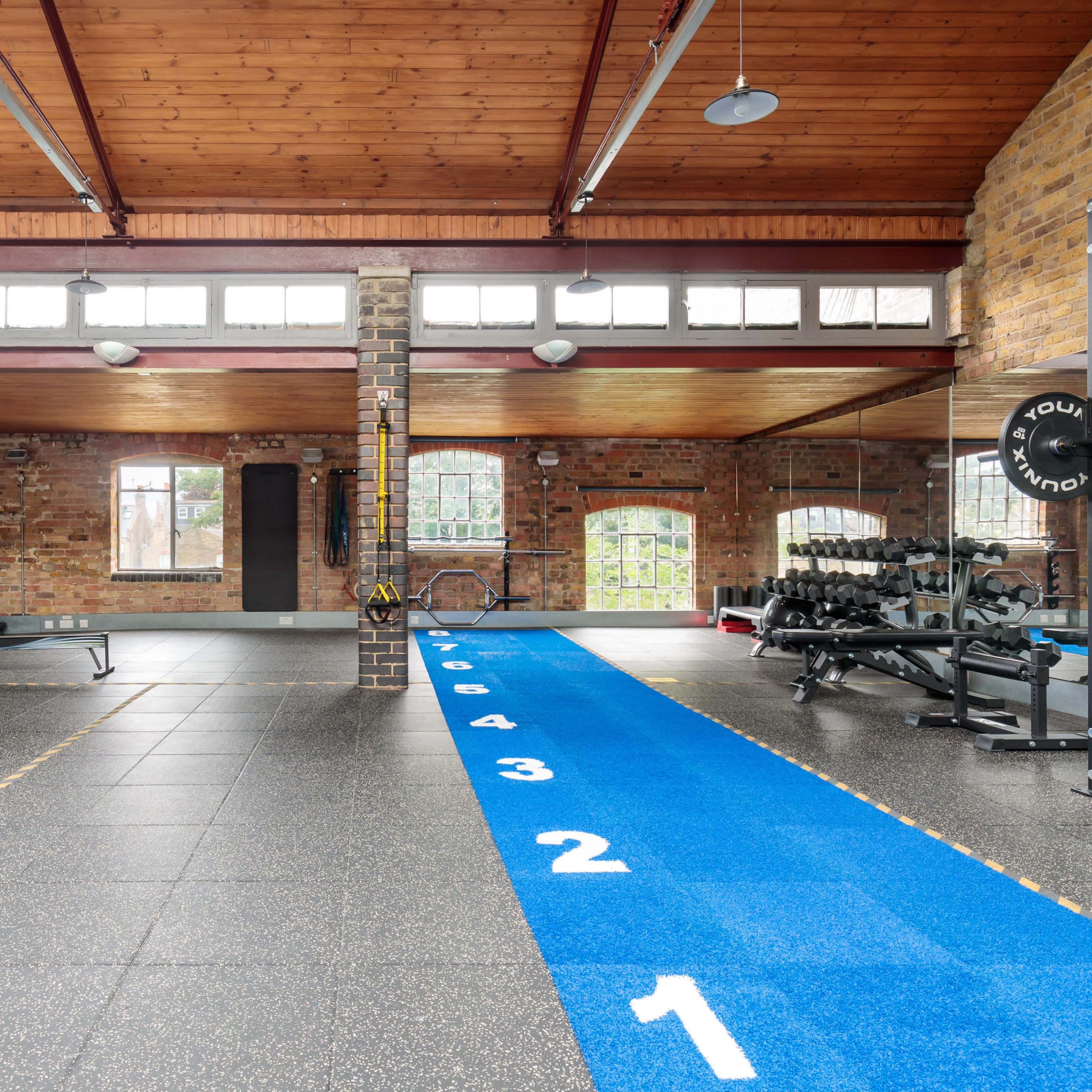 A spacious gym area featuring various workout equipment, a blue track numbered from one to eight, and large windows providing natural light.