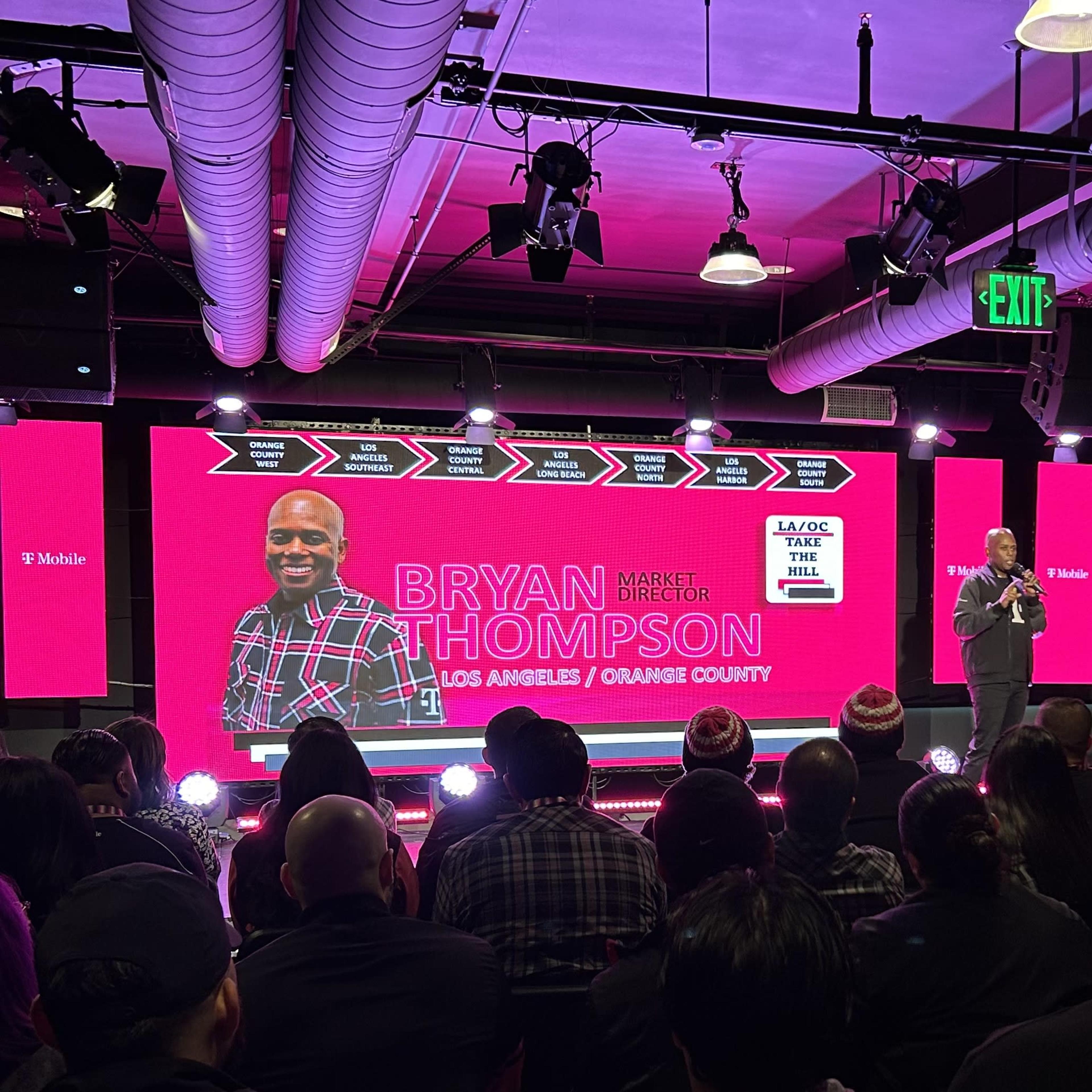 A speaker stands on stage in front of a large screen displaying their name and title during an event with a seated audience.