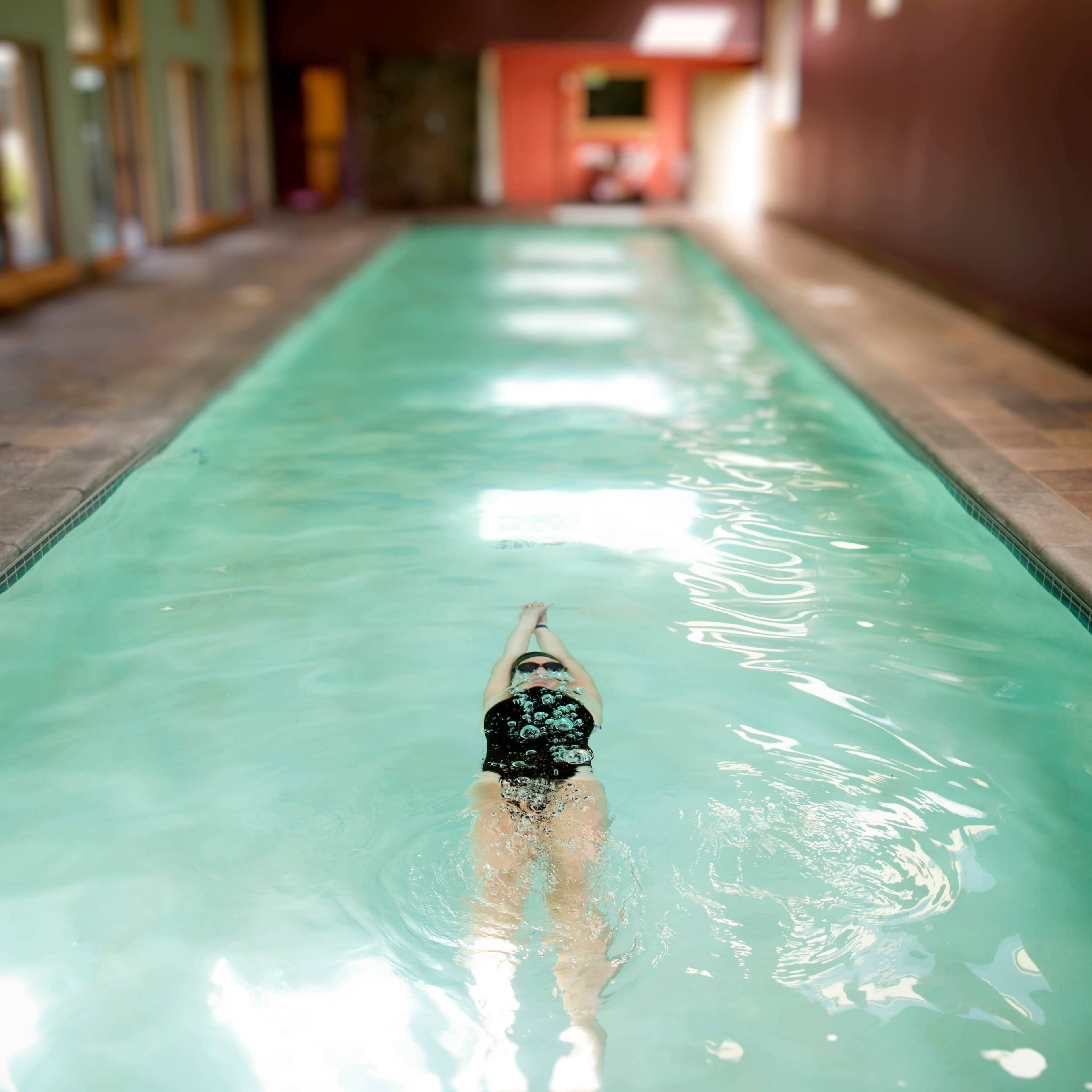 A swimmer in a black swimsuit glides through a narrow, indoor swimming pool lined with windows.