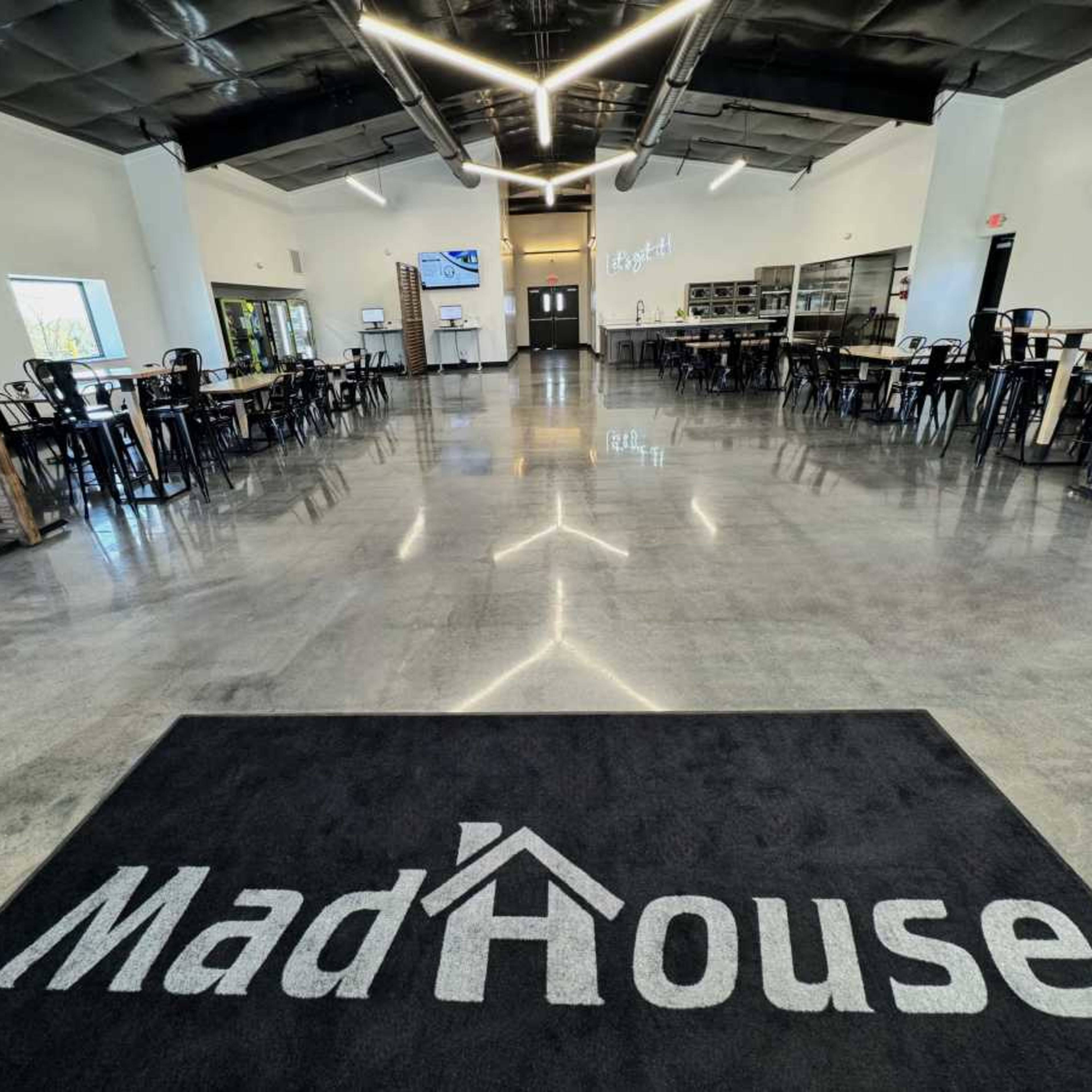 The interior of a modern dining space features a polished concrete floor, rows of black chairs and tables, and a prominent entrance rug with the name "MadHouse."