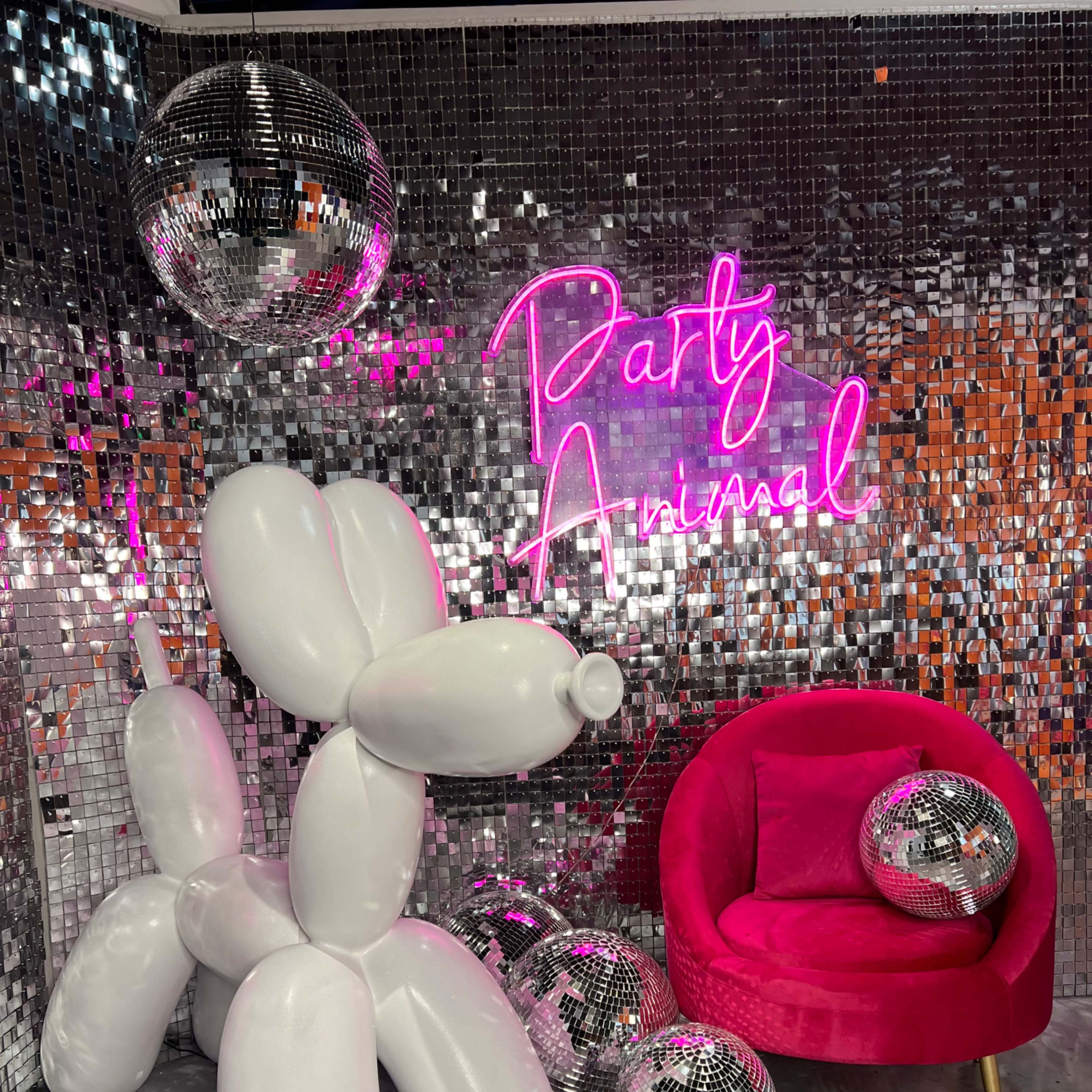 A white balloon dog stands next to a pink chair and scattered disco balls in front of a mirrored backdrop with a neon sign that reads "Party Animal."