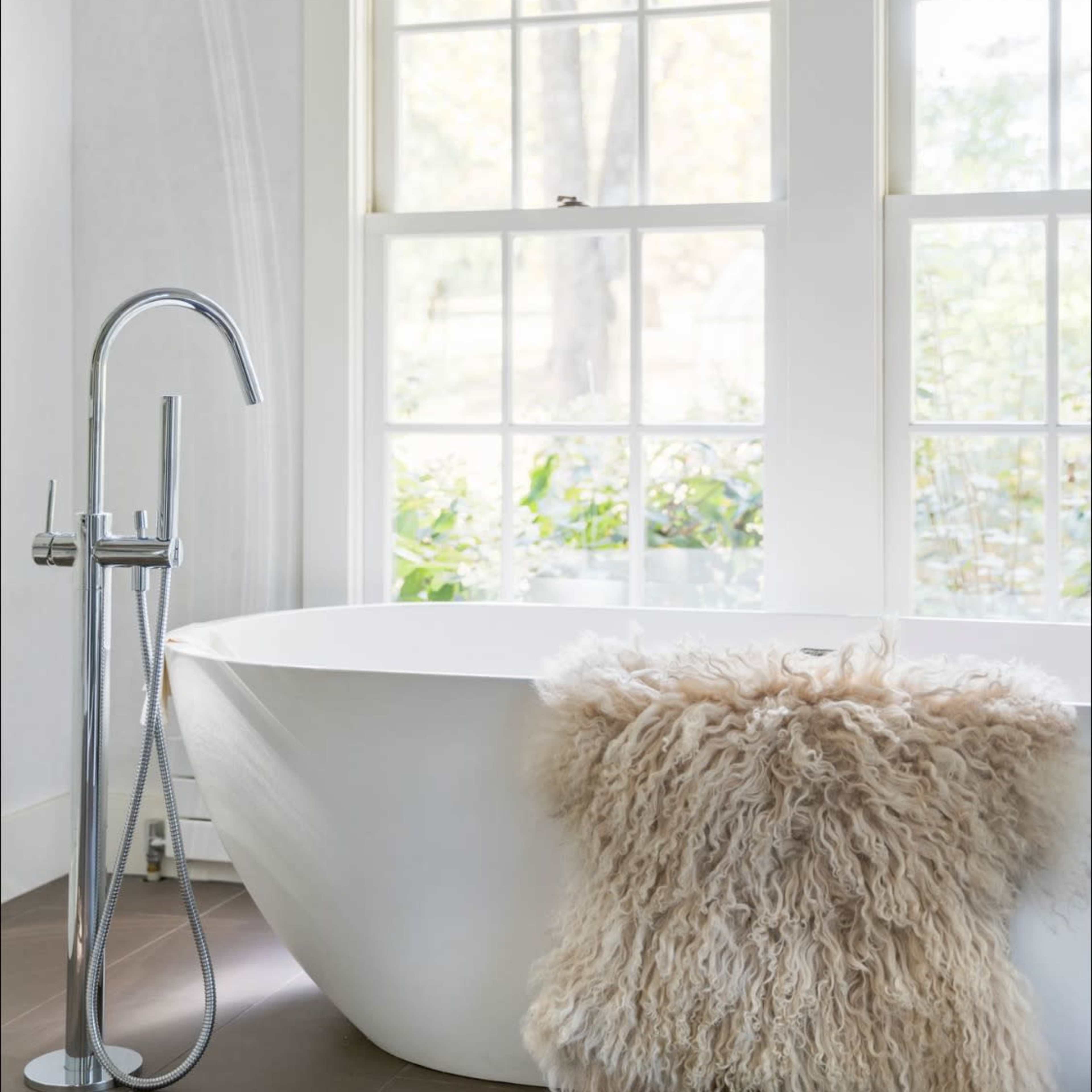 A modern bathroom features a freestanding bathtub beside a floor-mounted faucet, with a fluffy rug draped over its edge and large windows allowing natural light.