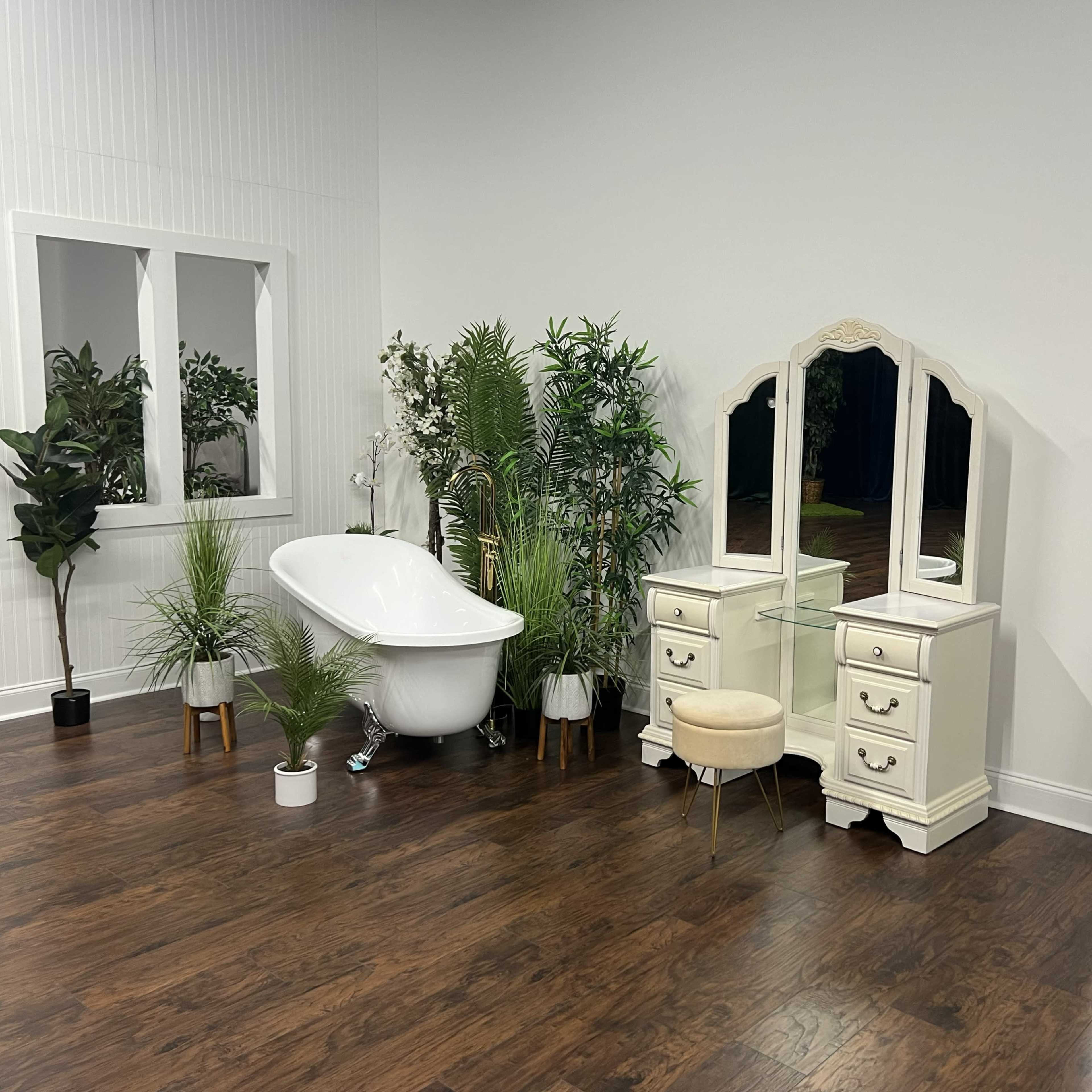 A minimalist bathroom setup featuring a freestanding bathtub, a vanity with two drawers, a stool, a large mirror, and various indoor plants.