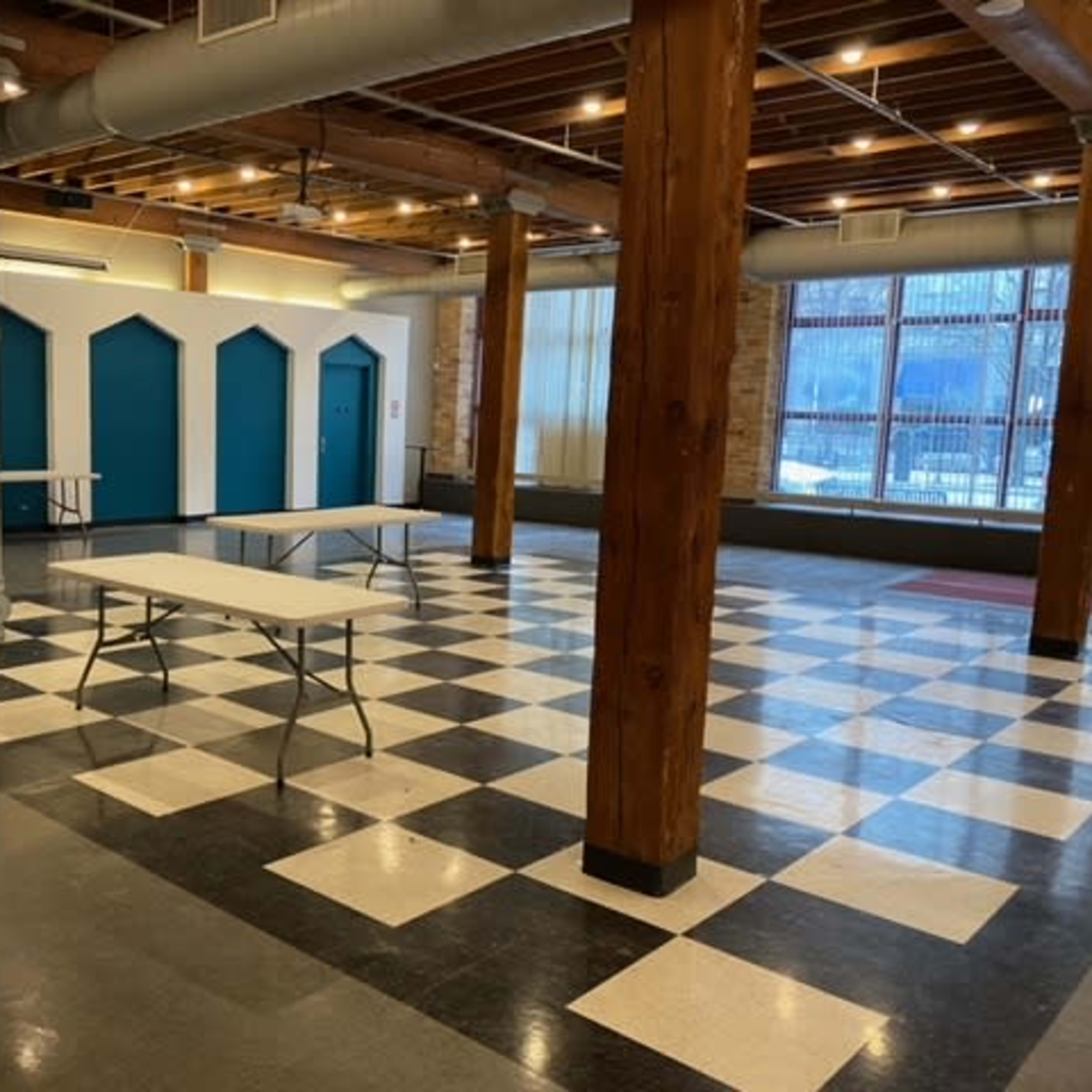 A large, open interior space with checkered flooring, wooden pillars, and several white folding tables arranged in front of teal doors.
