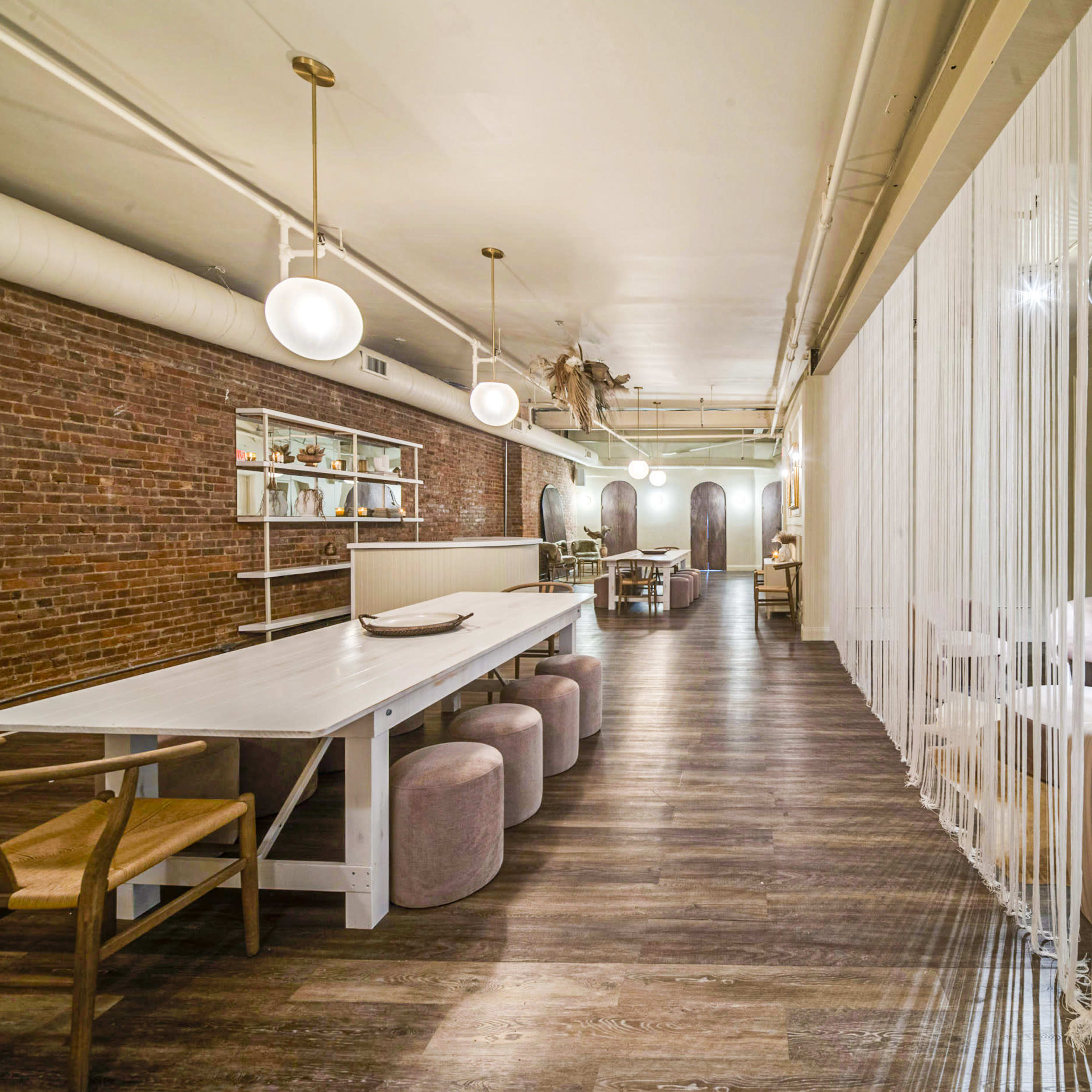A modern restaurant interior featuring a long white table, round stools, exposed brick walls, and dividers made of hanging threads.