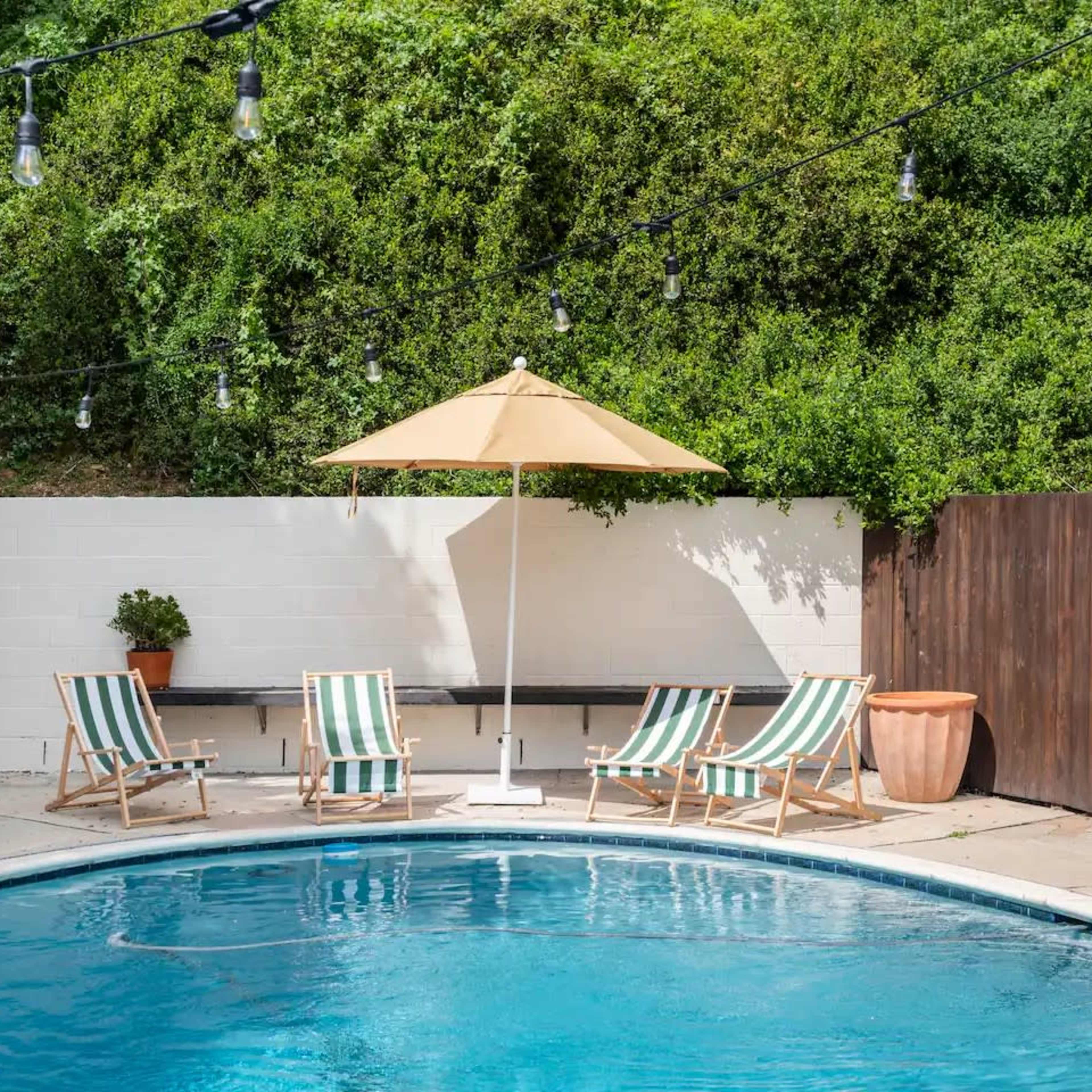 A swimming pool is surrounded by striped lounge chairs and an umbrella, with greenery and string lights in the background.