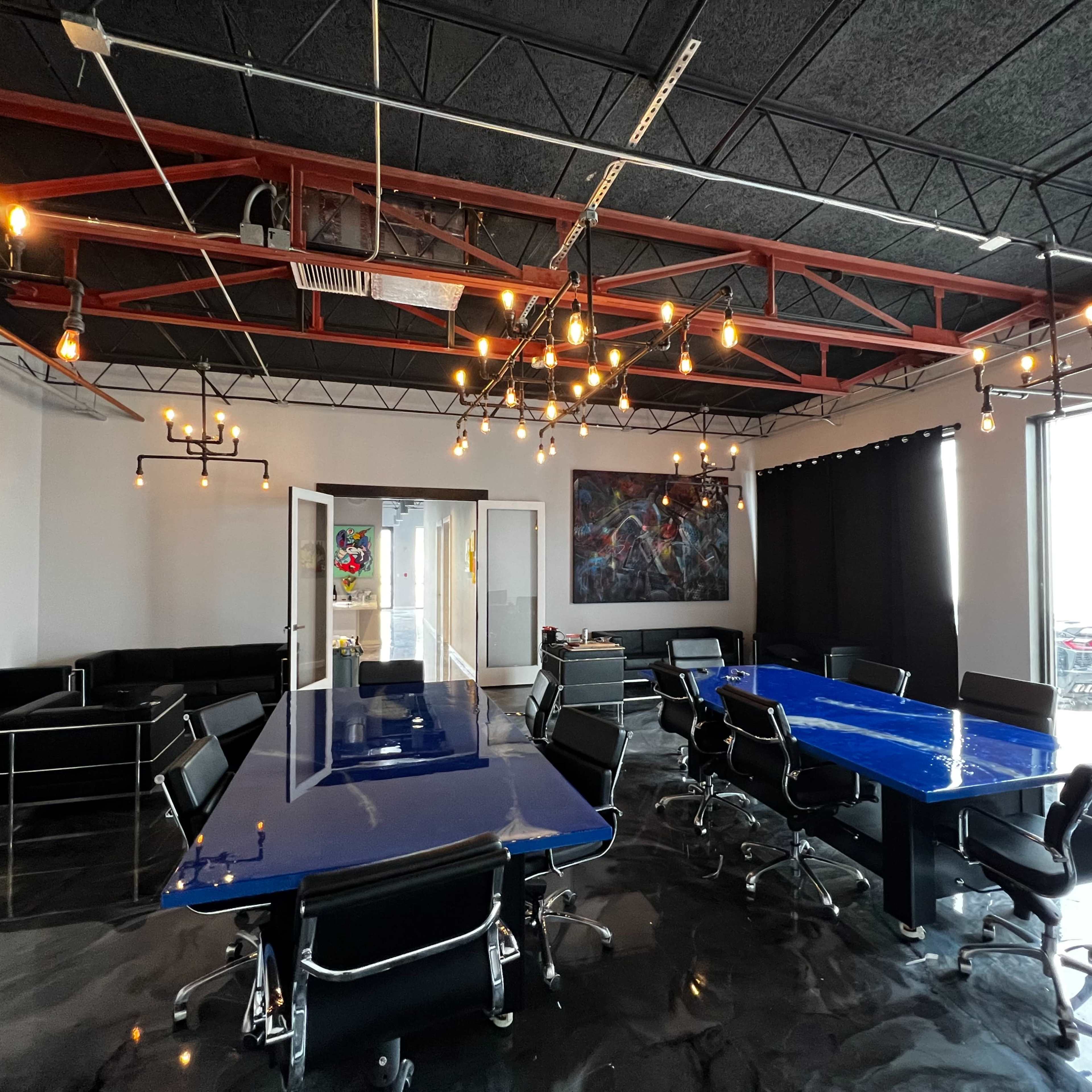 A modern conference room with a blue table, black chairs, exposed lighting, and large windows illuminating the space.