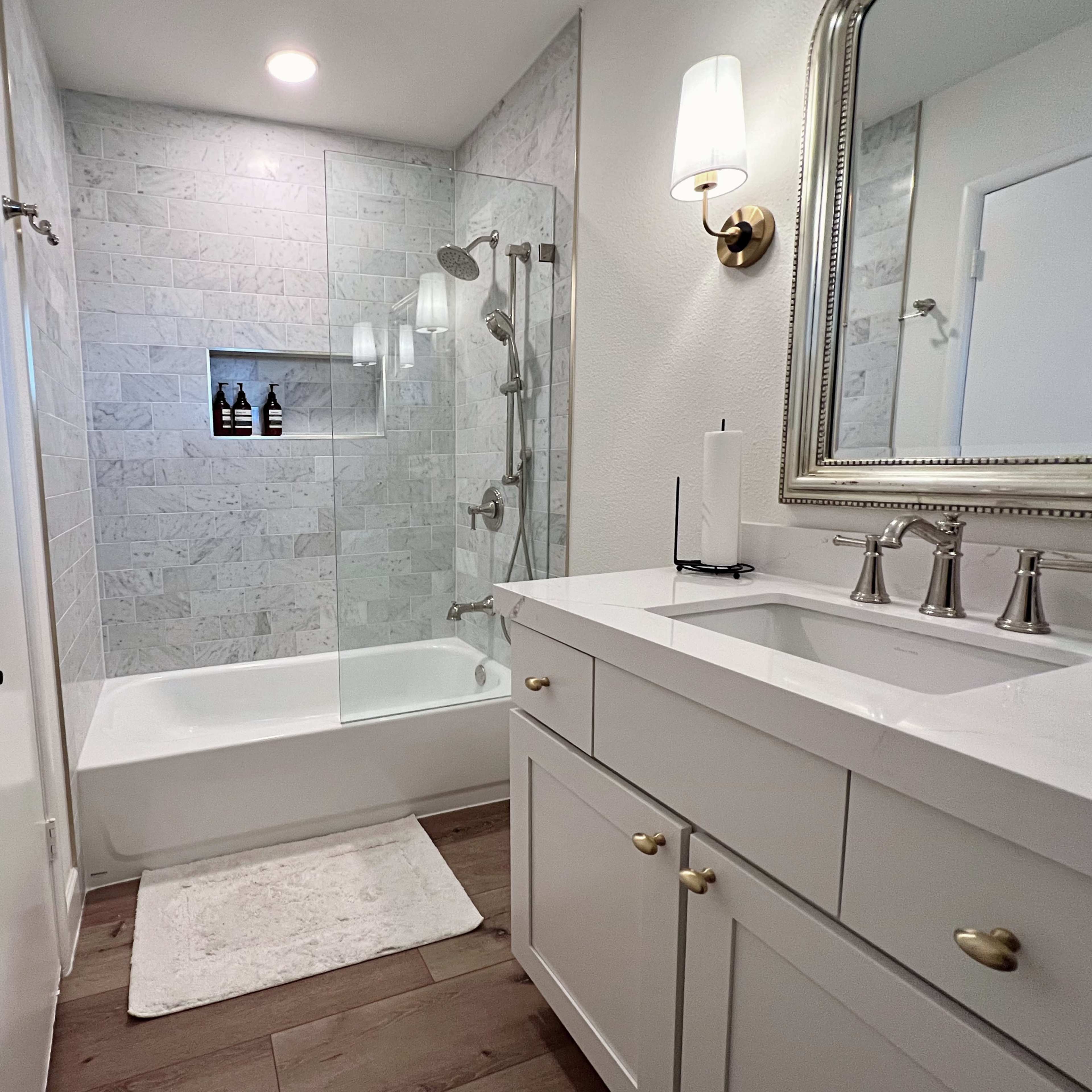 A modern bathroom featuring a glass-enclosed shower next to a white bathtub, a double-sink vanity with a mirror, and light-colored walls and flooring.