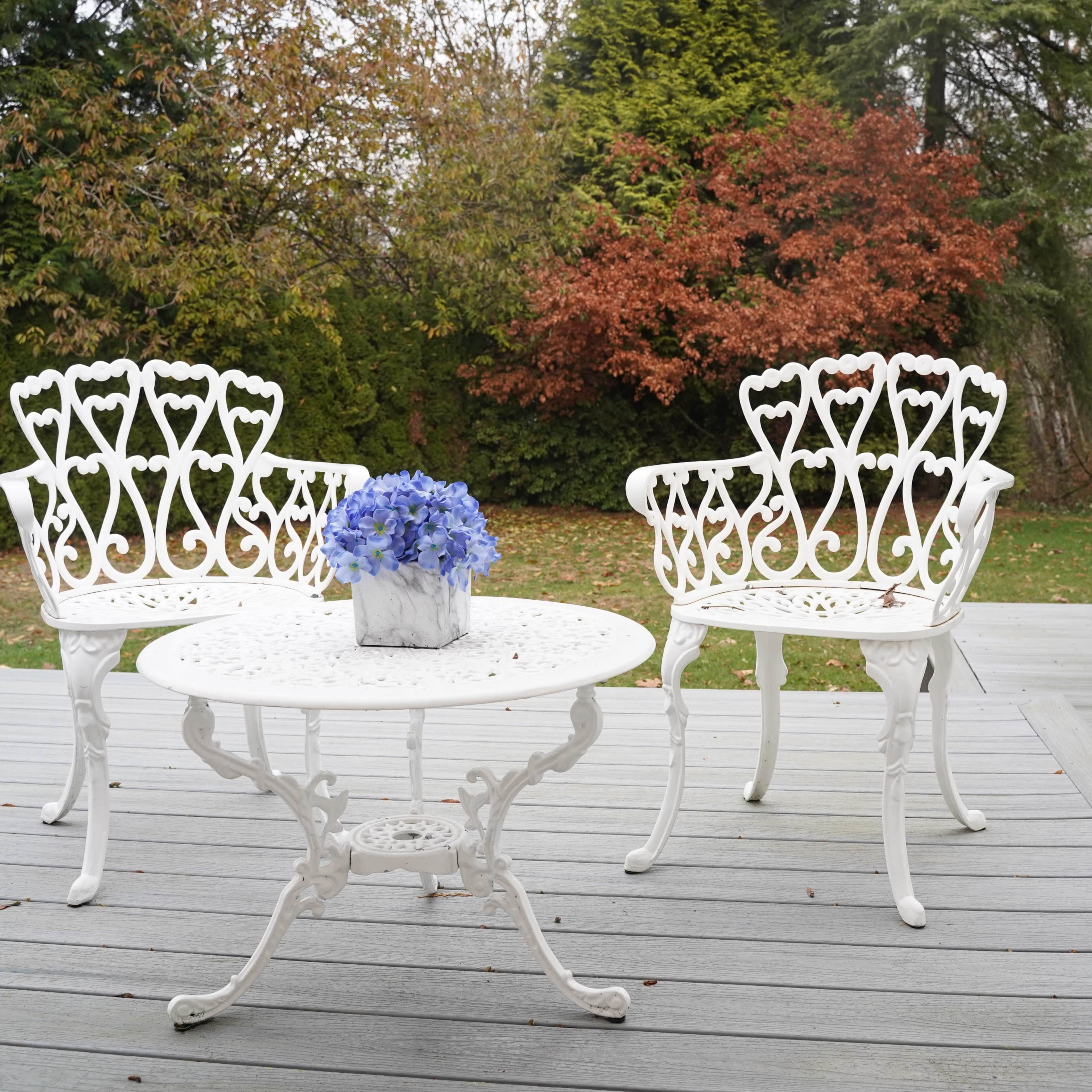 A small outdoor seating area features two white metal chairs and a round table with a vase of blue flowers, surrounded by greenery and autumn-colored trees.