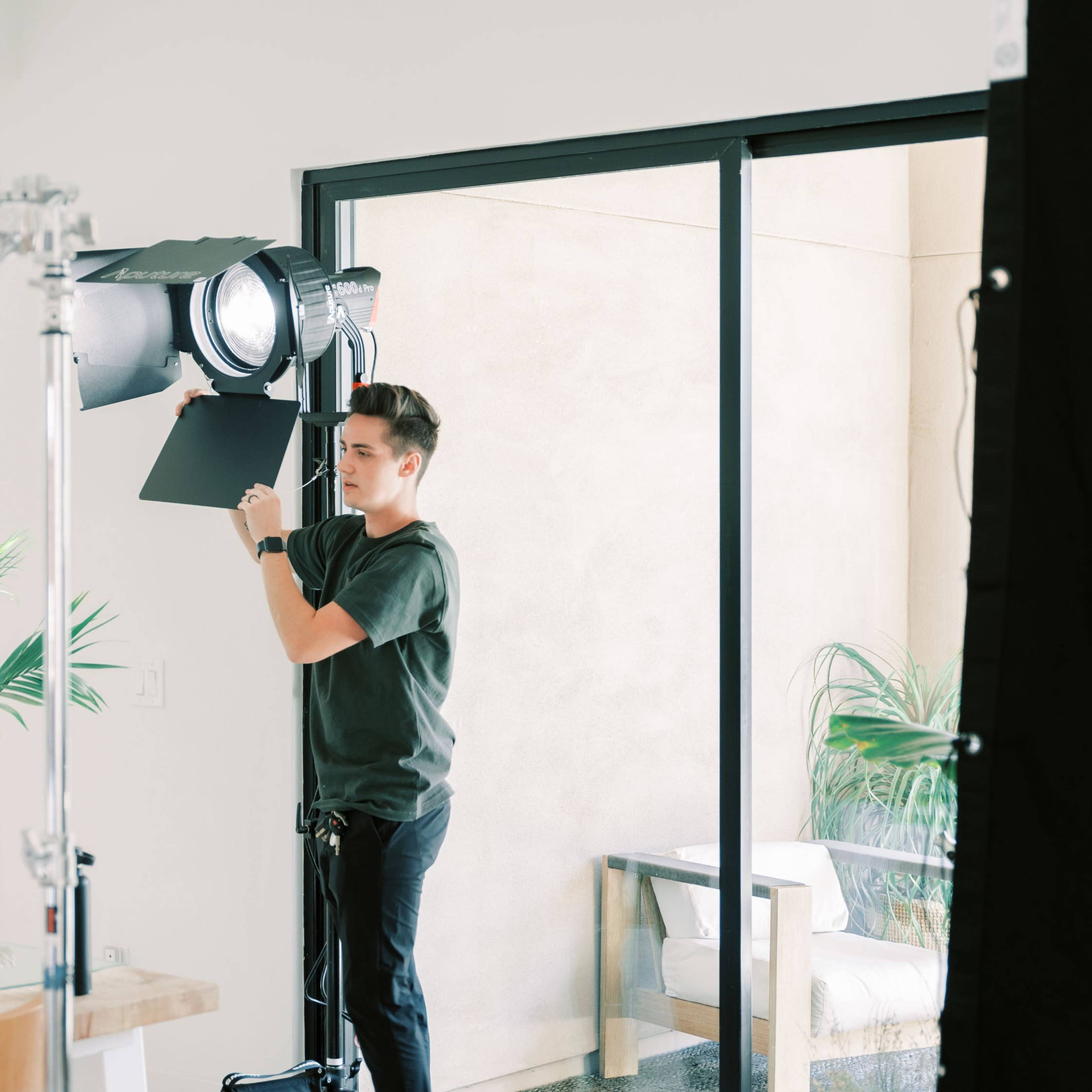A person adjusts a studio light in a bright room with large windows and indoor plants.