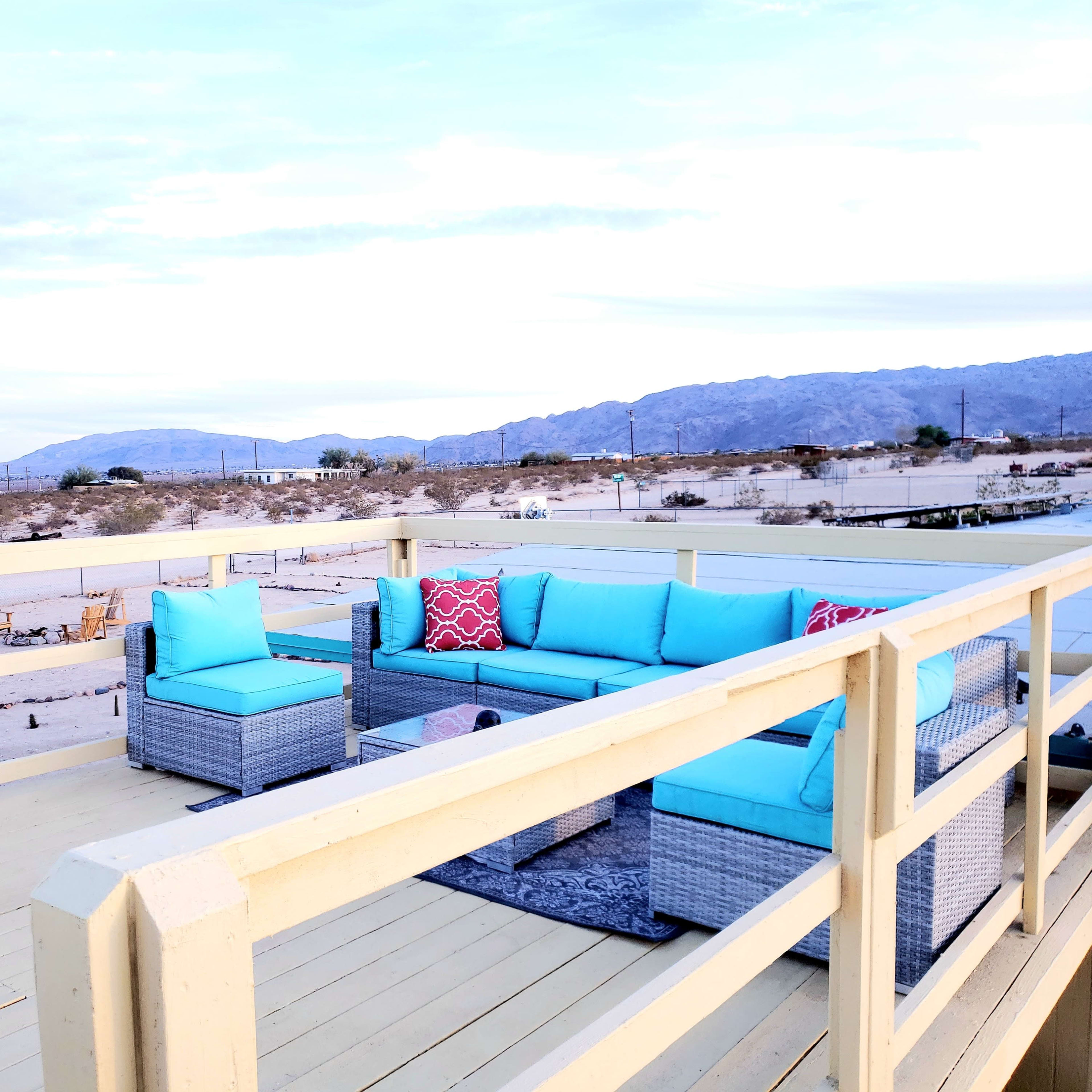 A turquoise outdoor seating area is positioned on a wooden deck overlooking a desert landscape with mountains in the background.