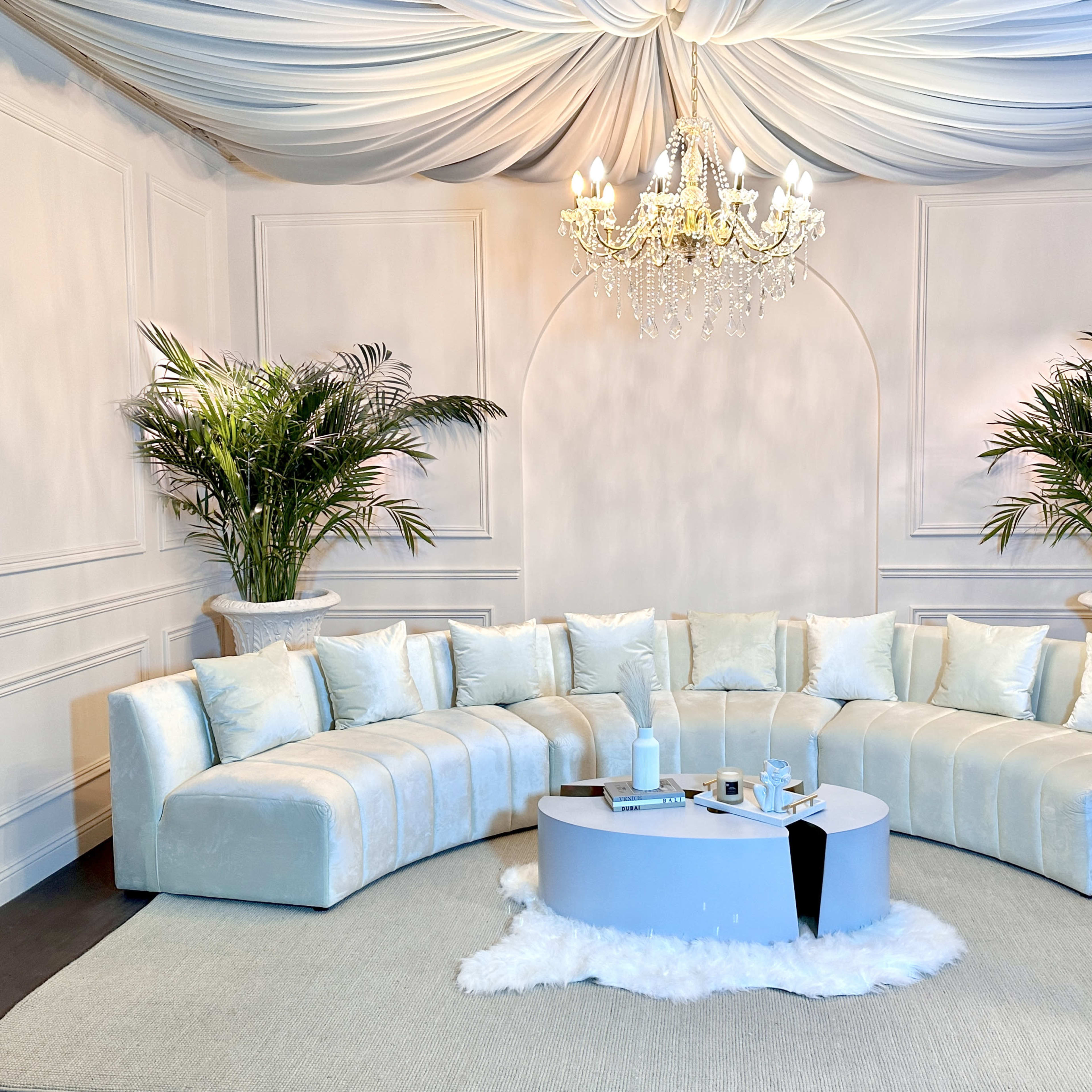 A spacious, elegantly decorated room featuring a curved white sofa, a round coffee table, two large potted plants, and a chandelier hanging from a draped ceiling.