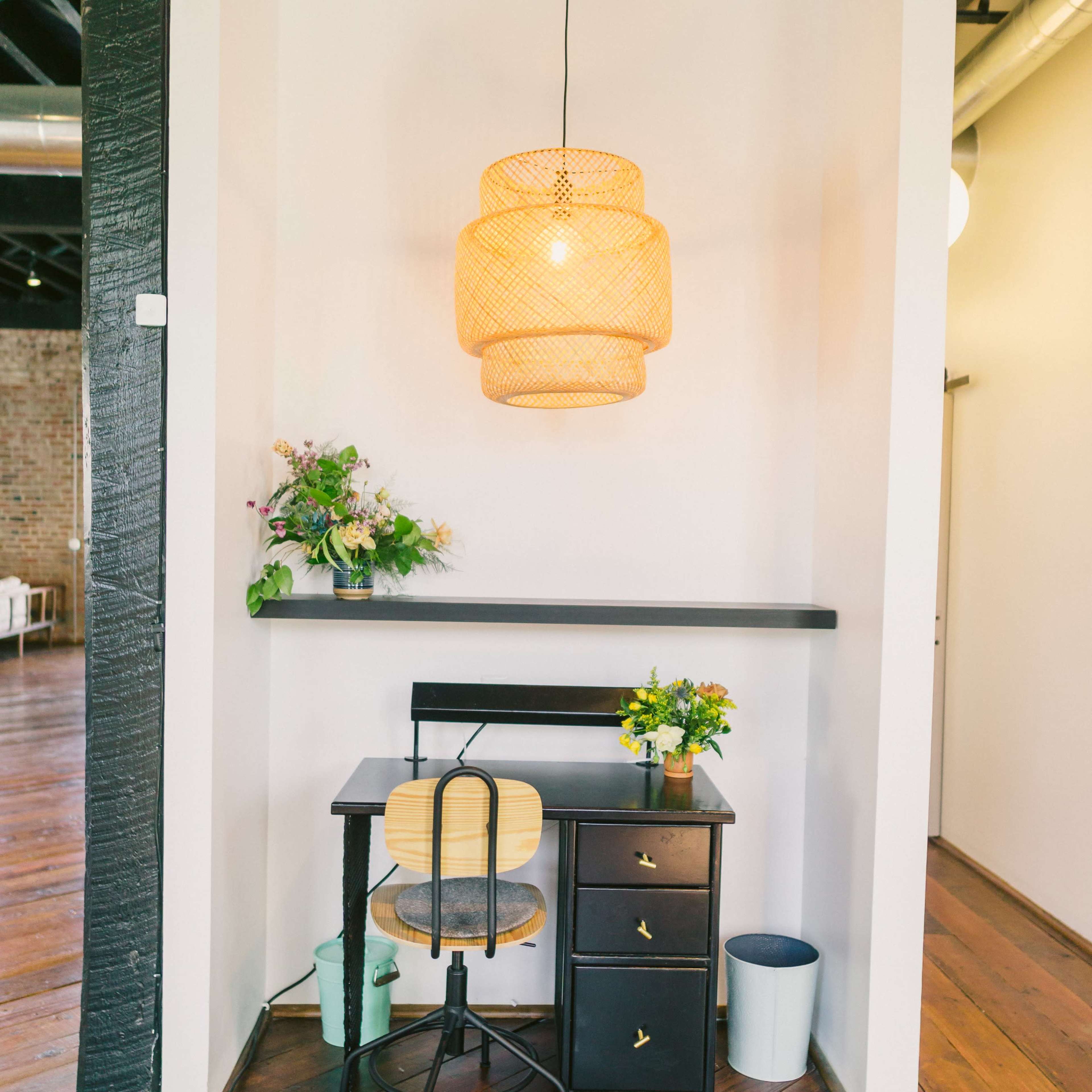 A small workspace features a black desk with a chair, a decorative lamp overhead, and a vase of flowers on the desk in a well-lit room with wooden flooring.