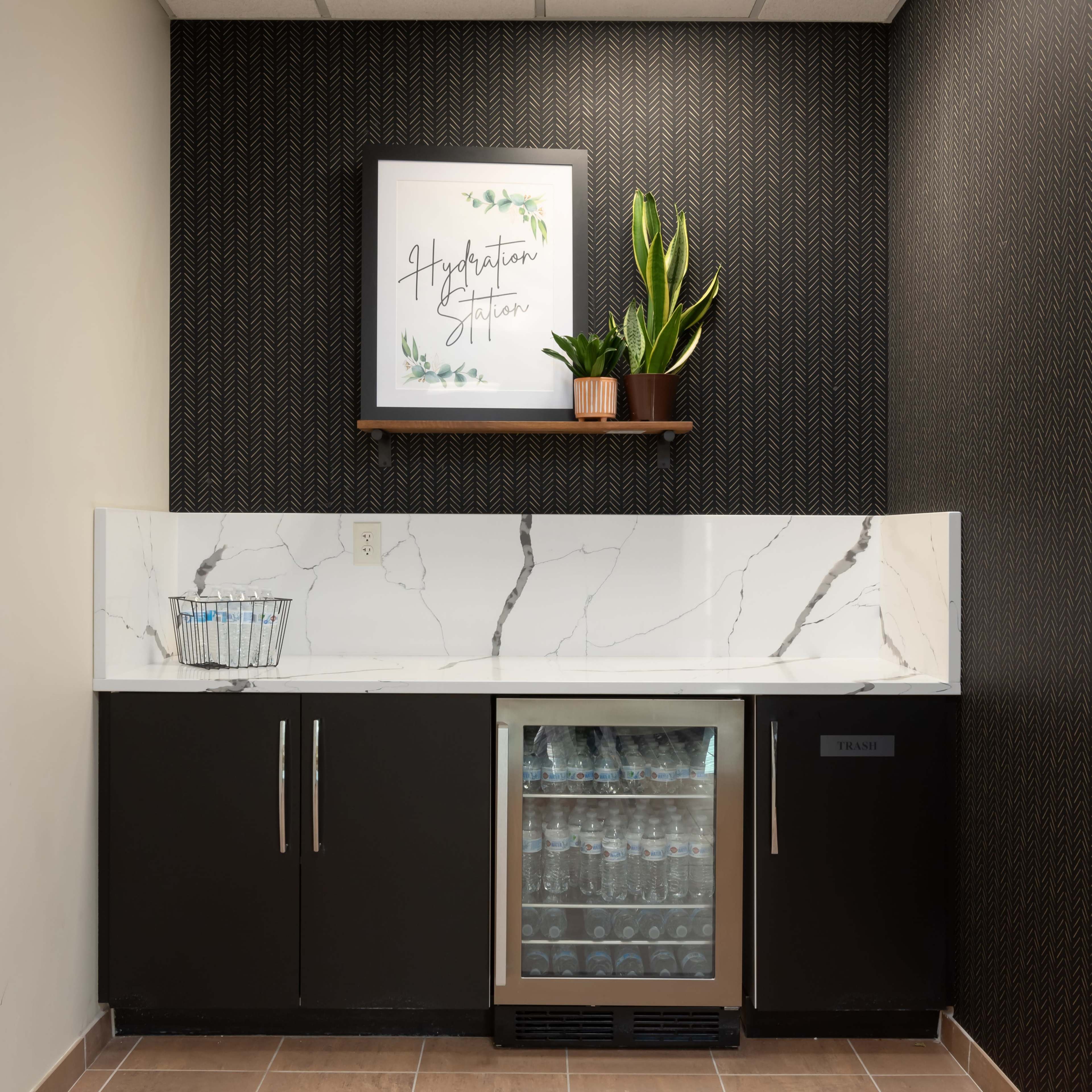 A hydration station with a marble countertop, a plant on a shelf, and a mini refrigerator beneath for beverages.