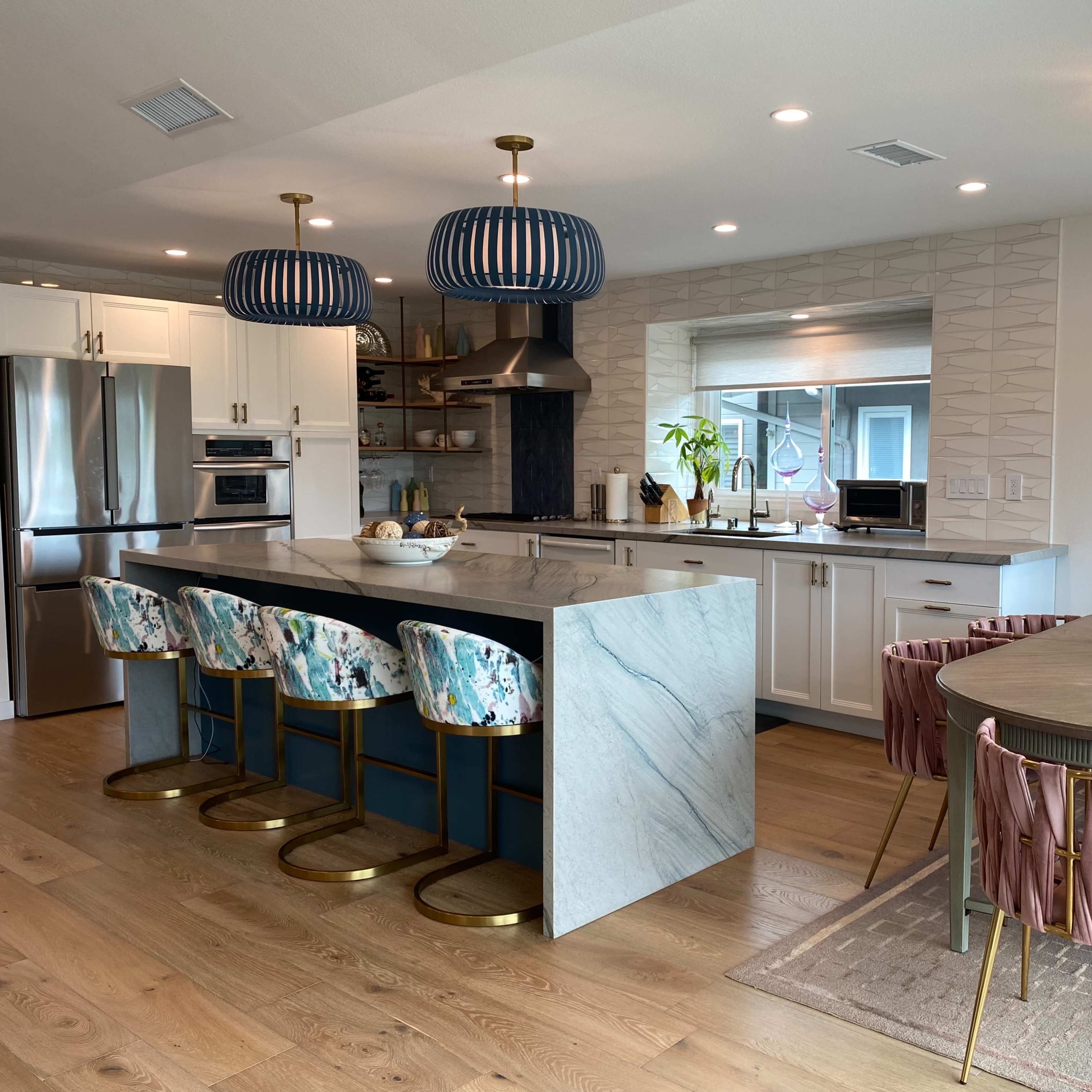 A modern kitchen featuring a marble island with four stools, a dining table with pink and green chairs, and decorative pendant lights.