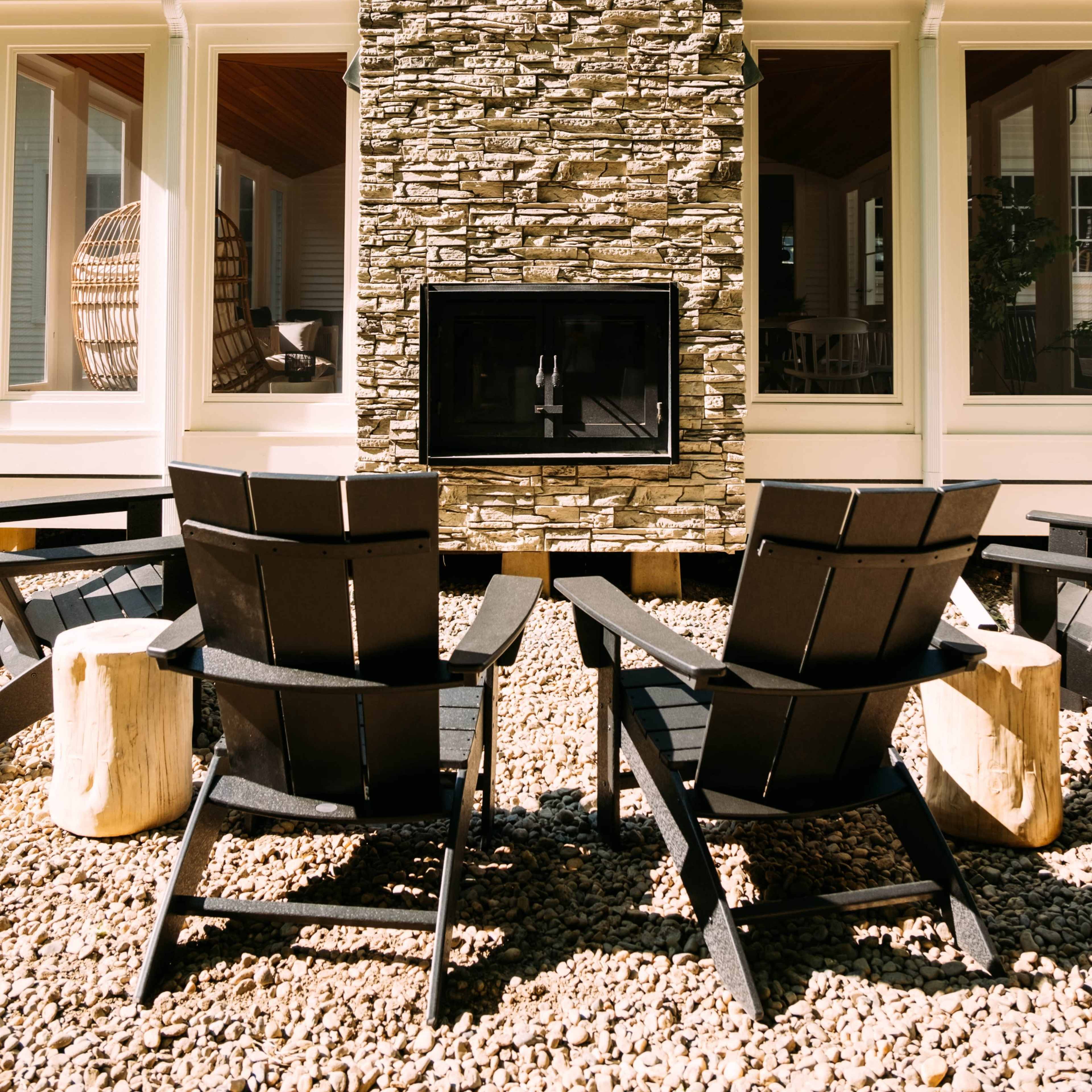 Two black adirondack chairs face a stone fireplace in a courtyard surrounded by large windows.