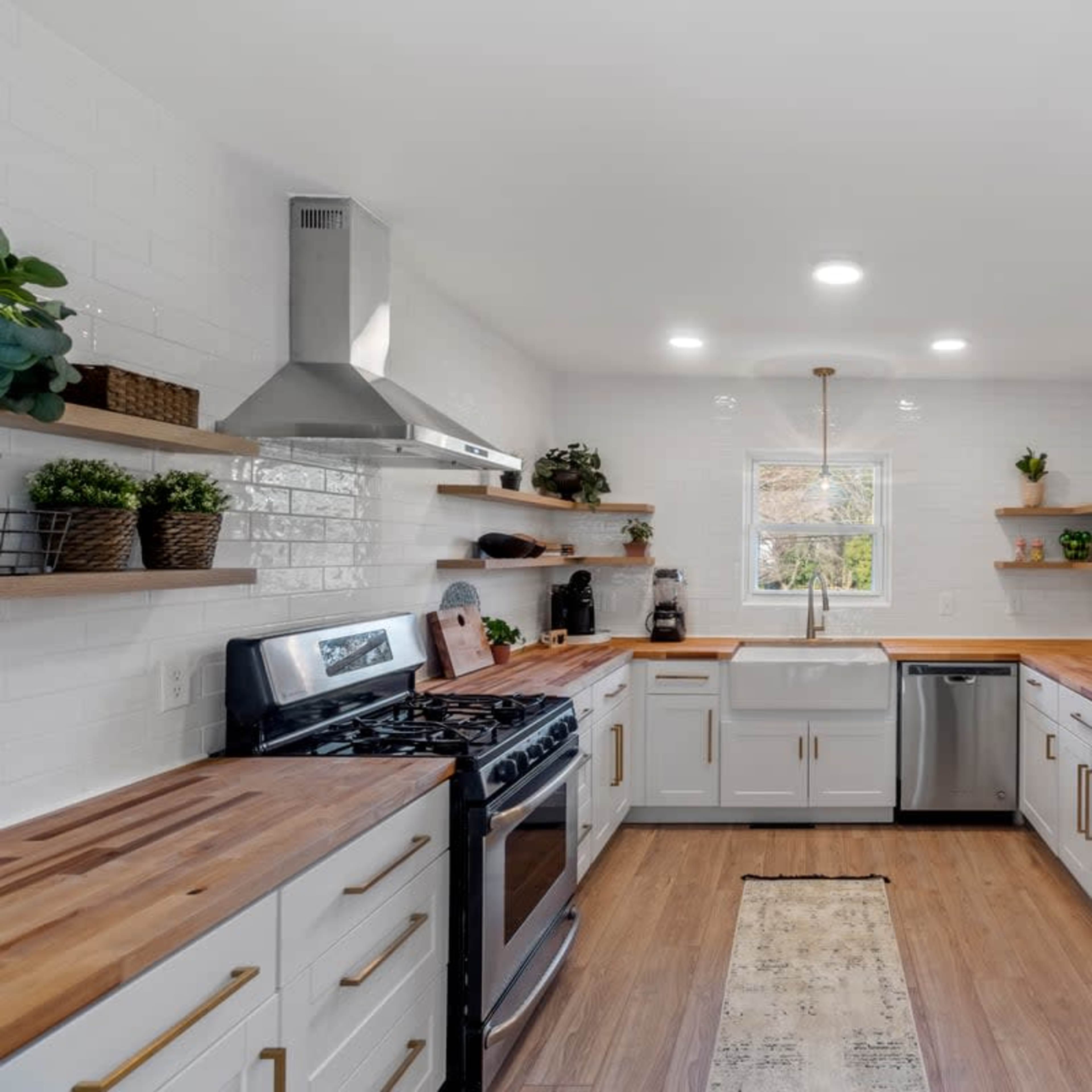A modern kitchen with stainless steel appliances, white cabinetry, wooden countertops, and open shelving with plants and kitchen items.