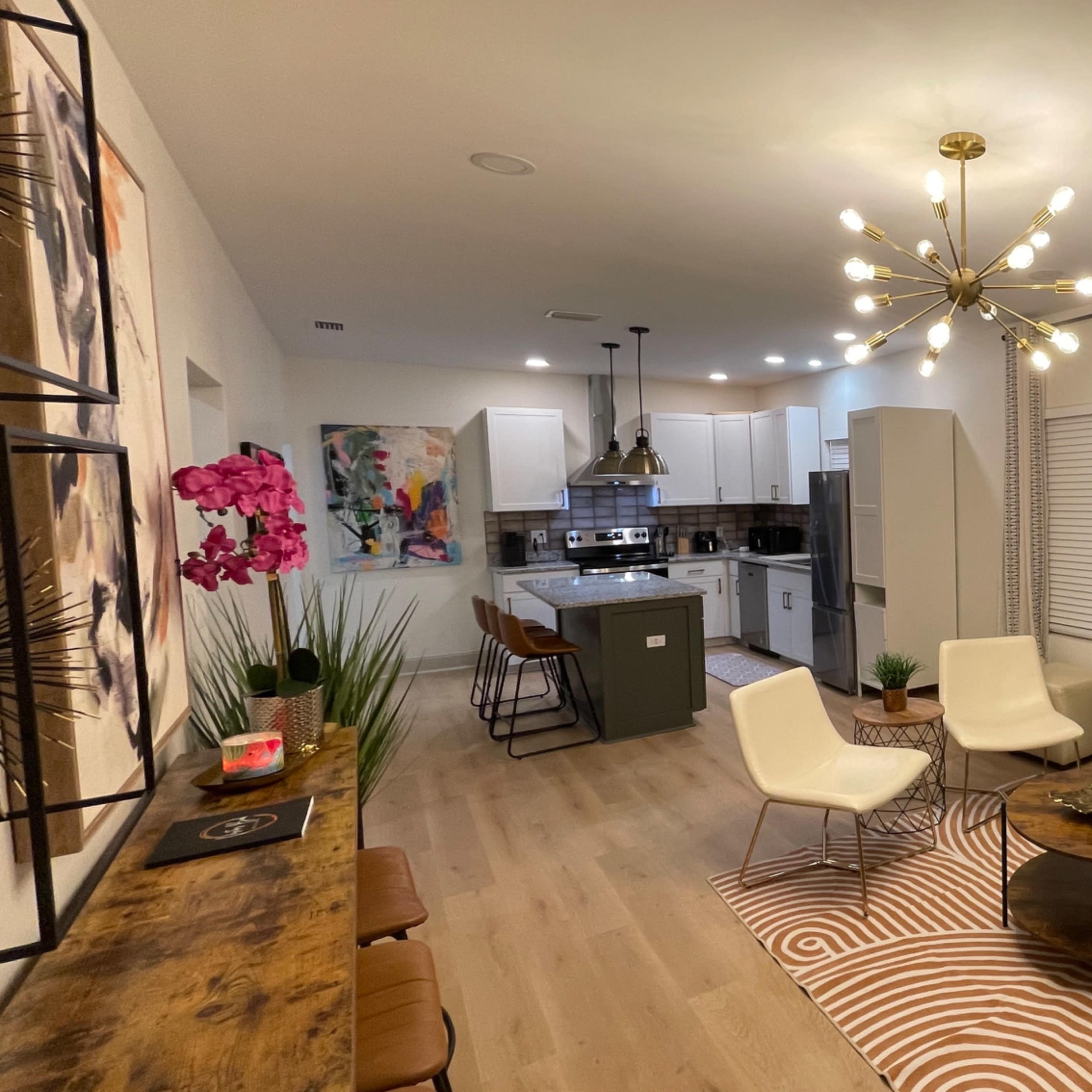 A modern living room with a brown leather sofa, a circular coffee table, and a wall-mounted faux fireplace, connected to an open kitchen with a large island.