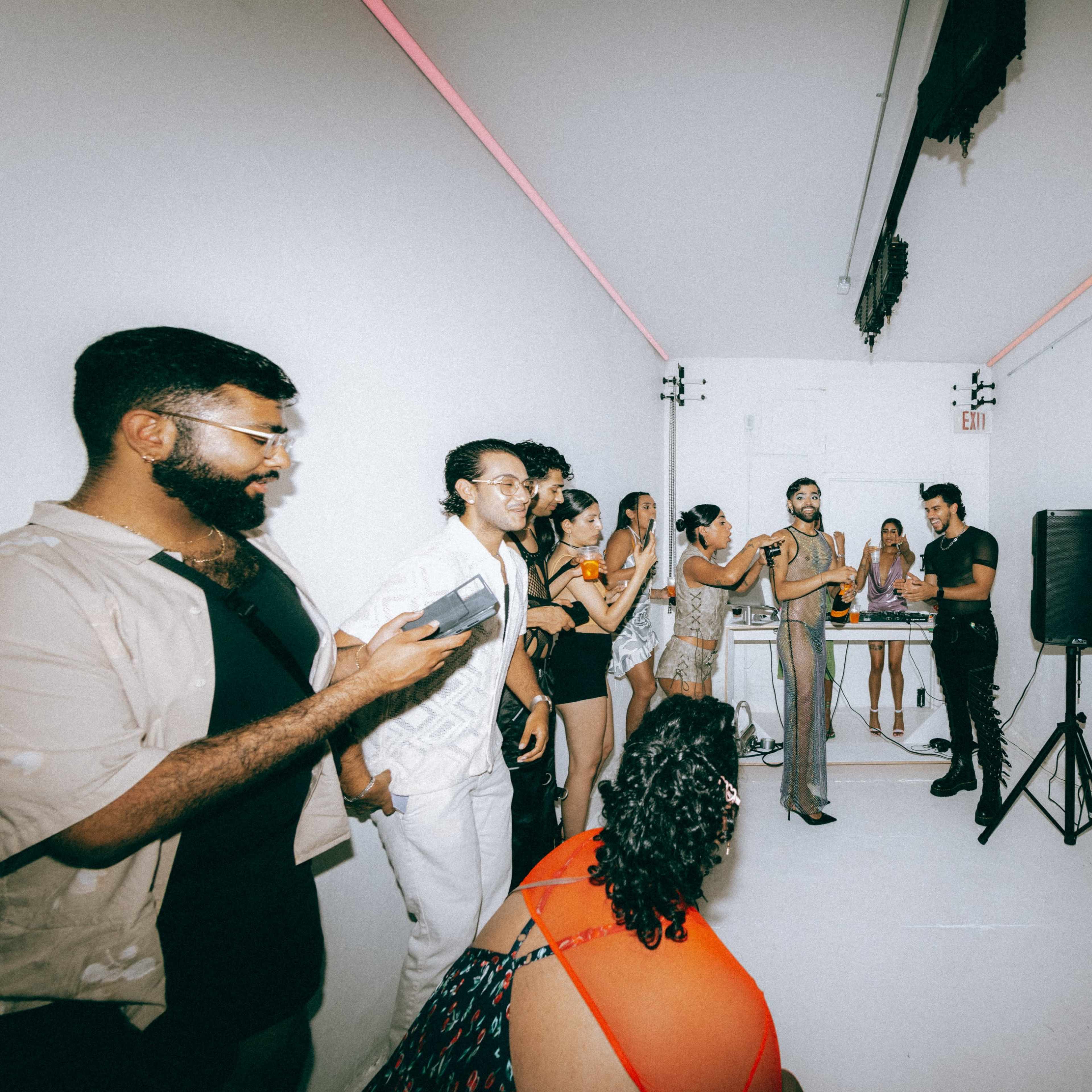 A group of people interacts in a white room with a DJ setup and colorful lighting, while one person stands at a microphone.