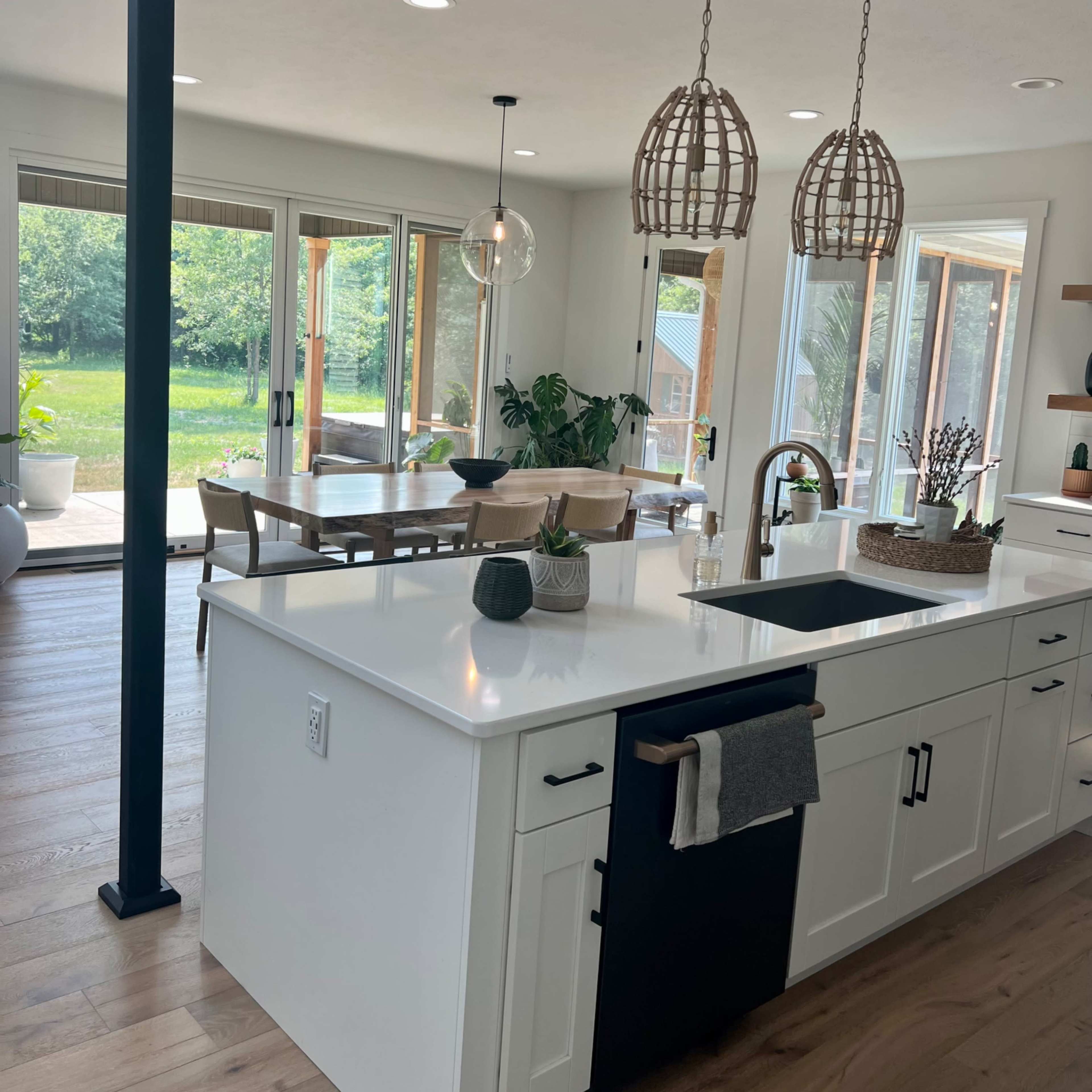 A modern kitchen features a central island with a sink, black cabinetry, and two decorative pendant lights, overlooking a dining area and large windows that provide views of the backyard.
