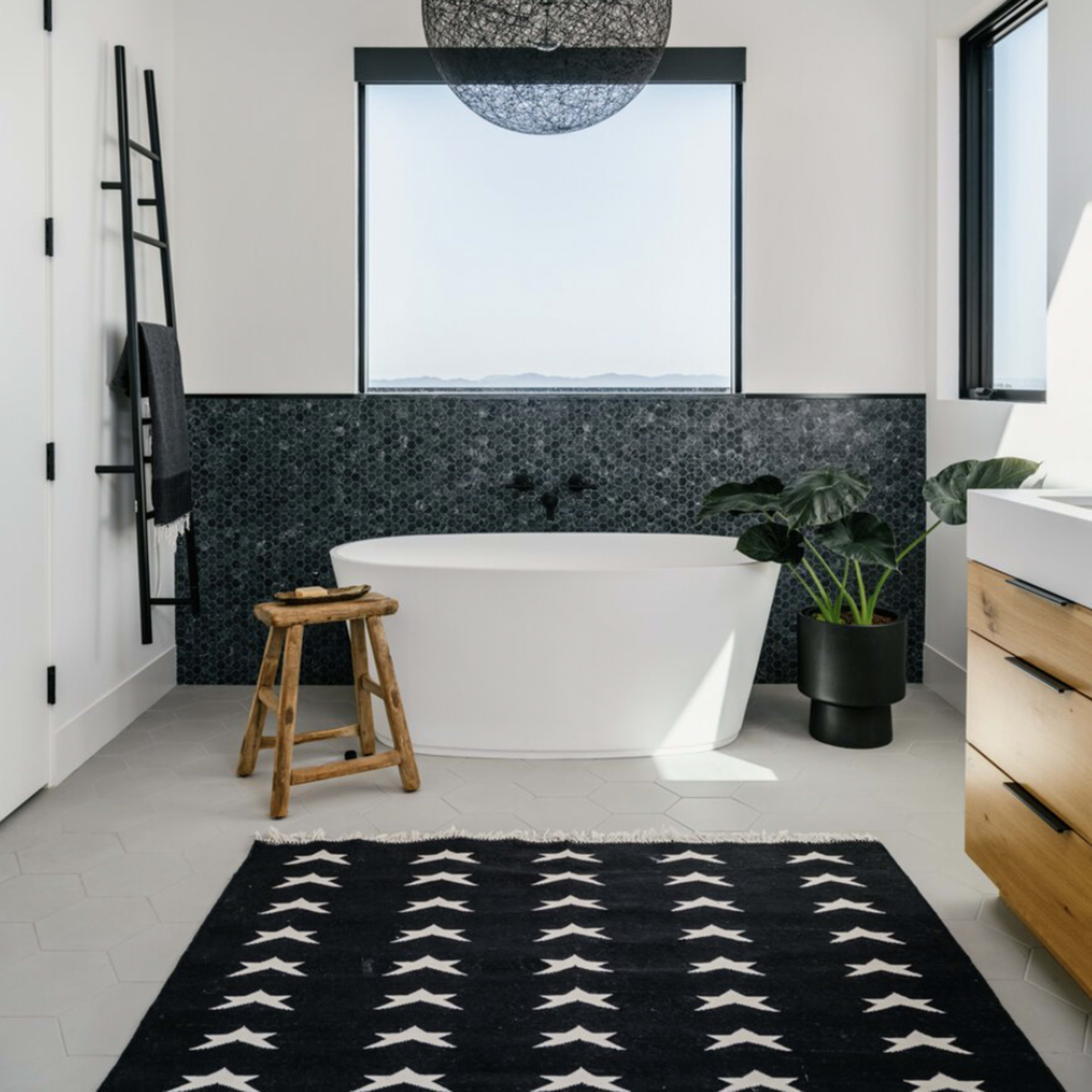 A modern bathroom features a freestanding bathtub, large window with a scenic view, and a patterned rug on the floor.