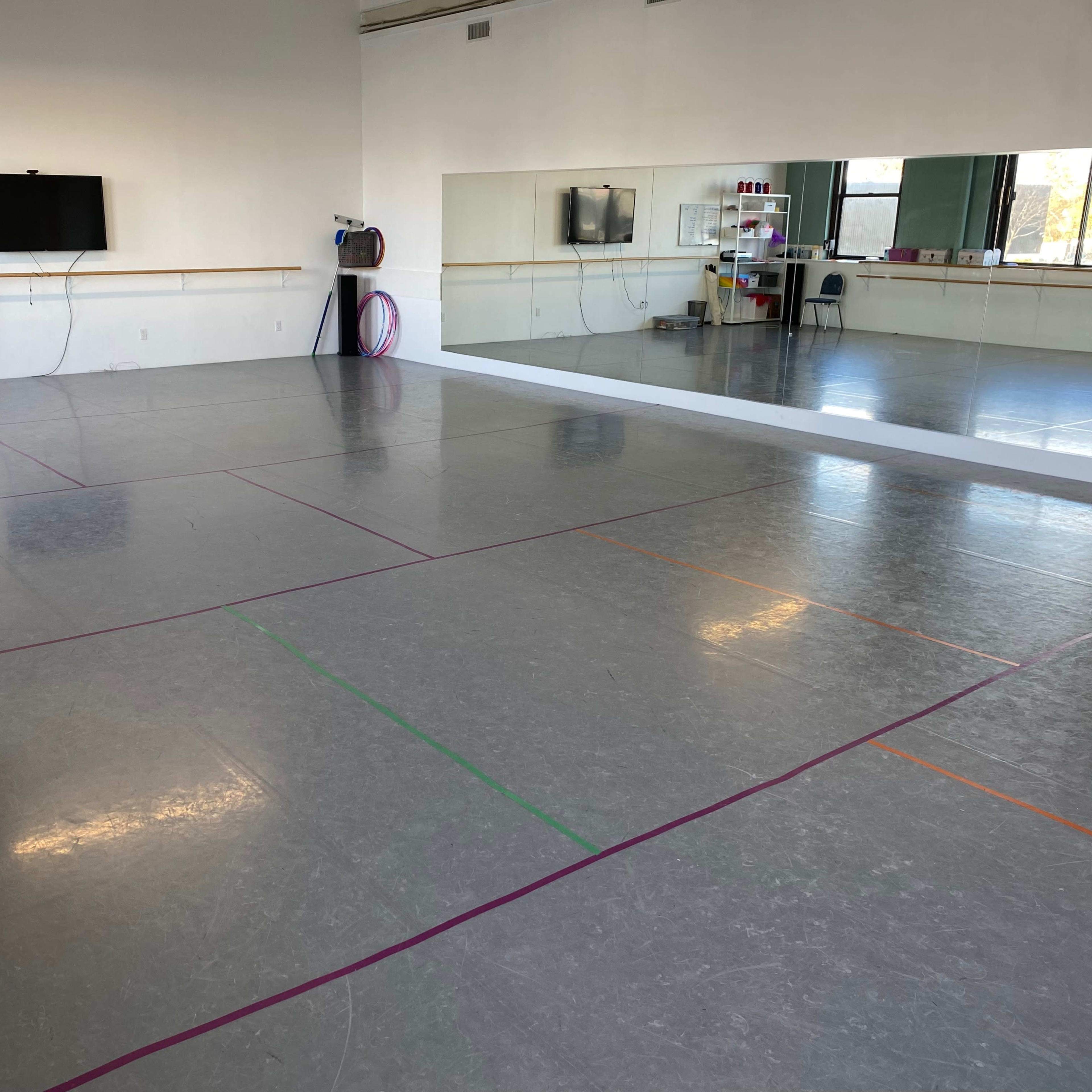 A spacious dance studio with a gray floor, wall mirrors, and minimal decor, featuring marked lines in various colors on the floor.