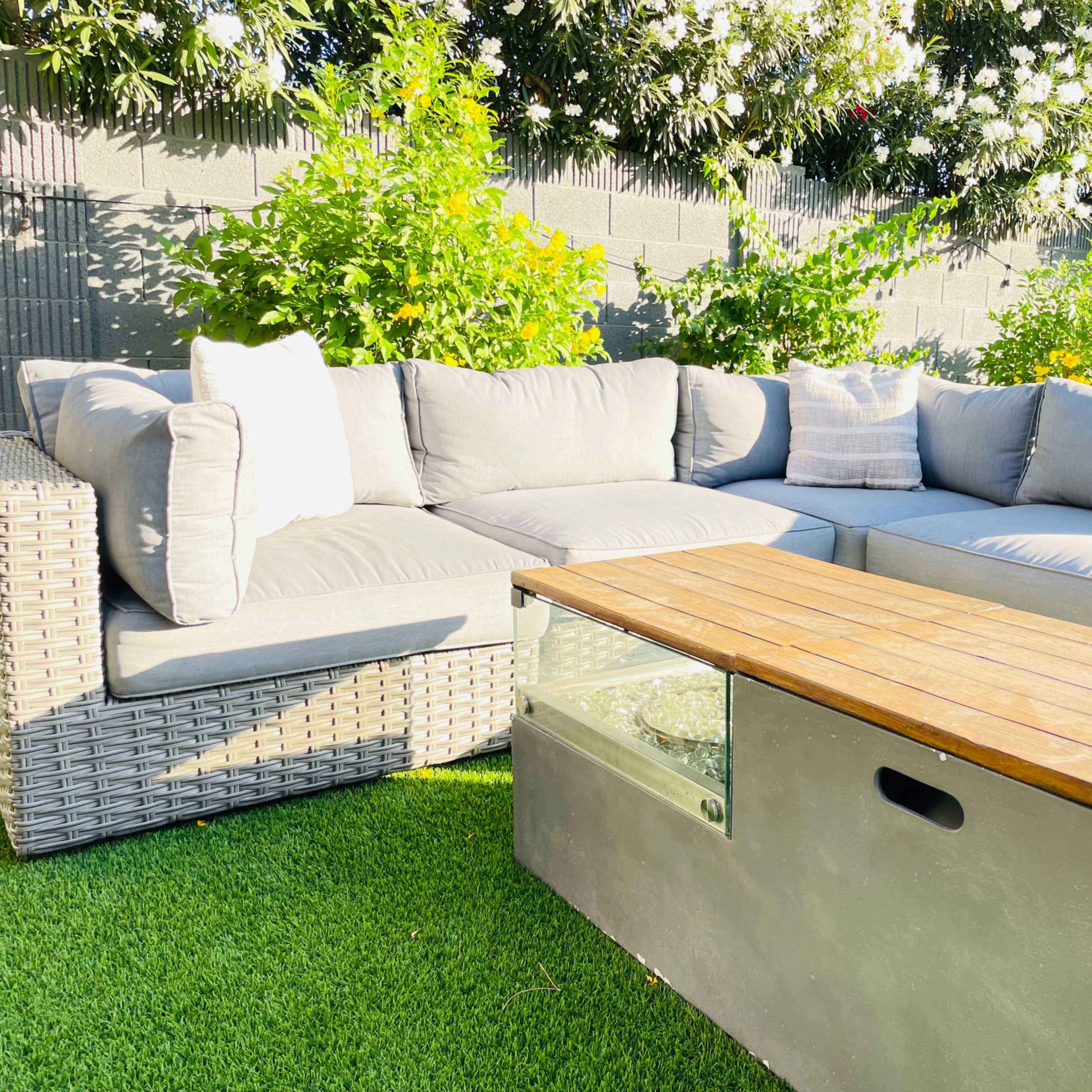 A sectional outdoor couch with gray cushions is positioned beside a wooden coffee table on a grassy area surrounded by flowering plants.