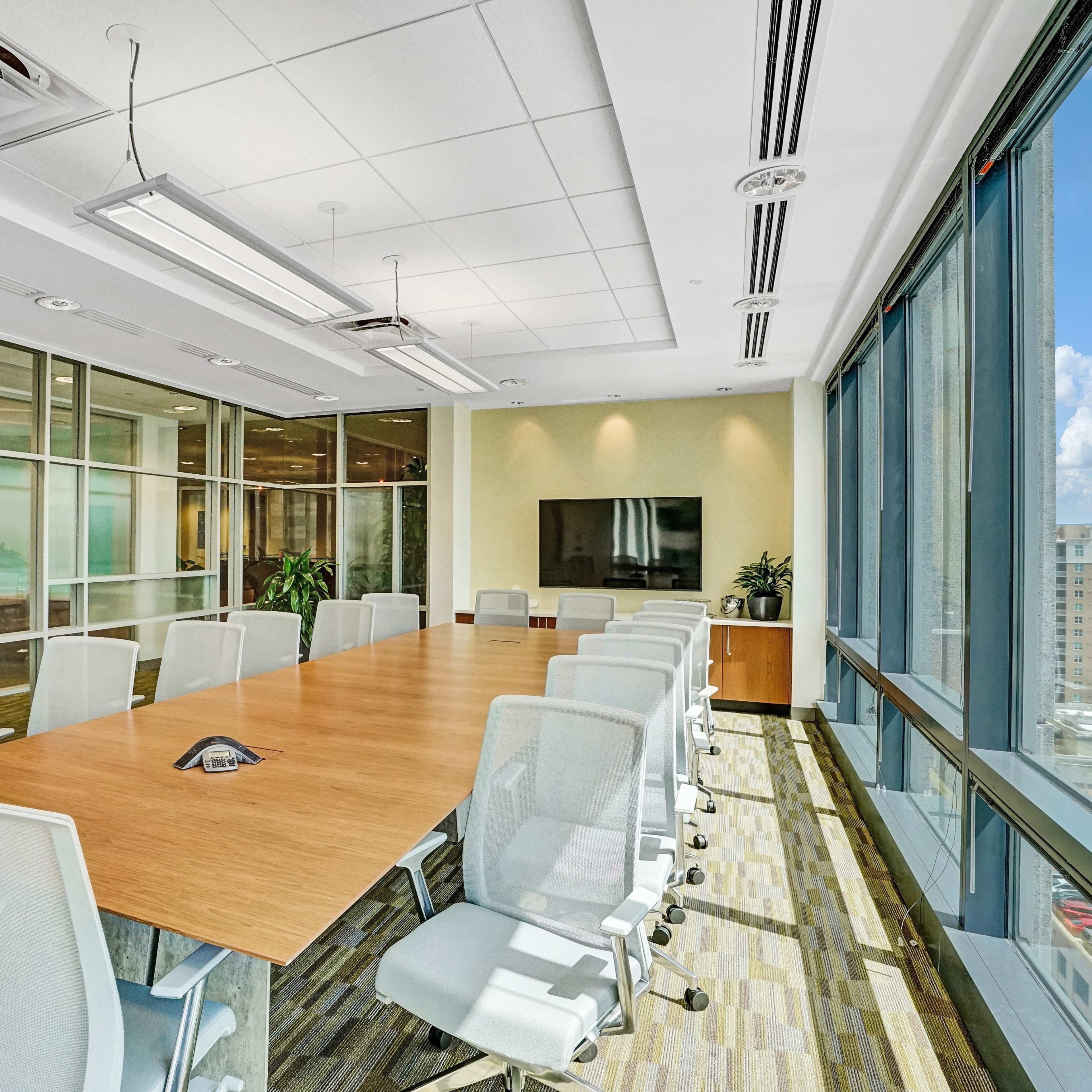 A modern conference room with a long wooden table surrounded by white chairs, large windows, and a wall-mounted TV.