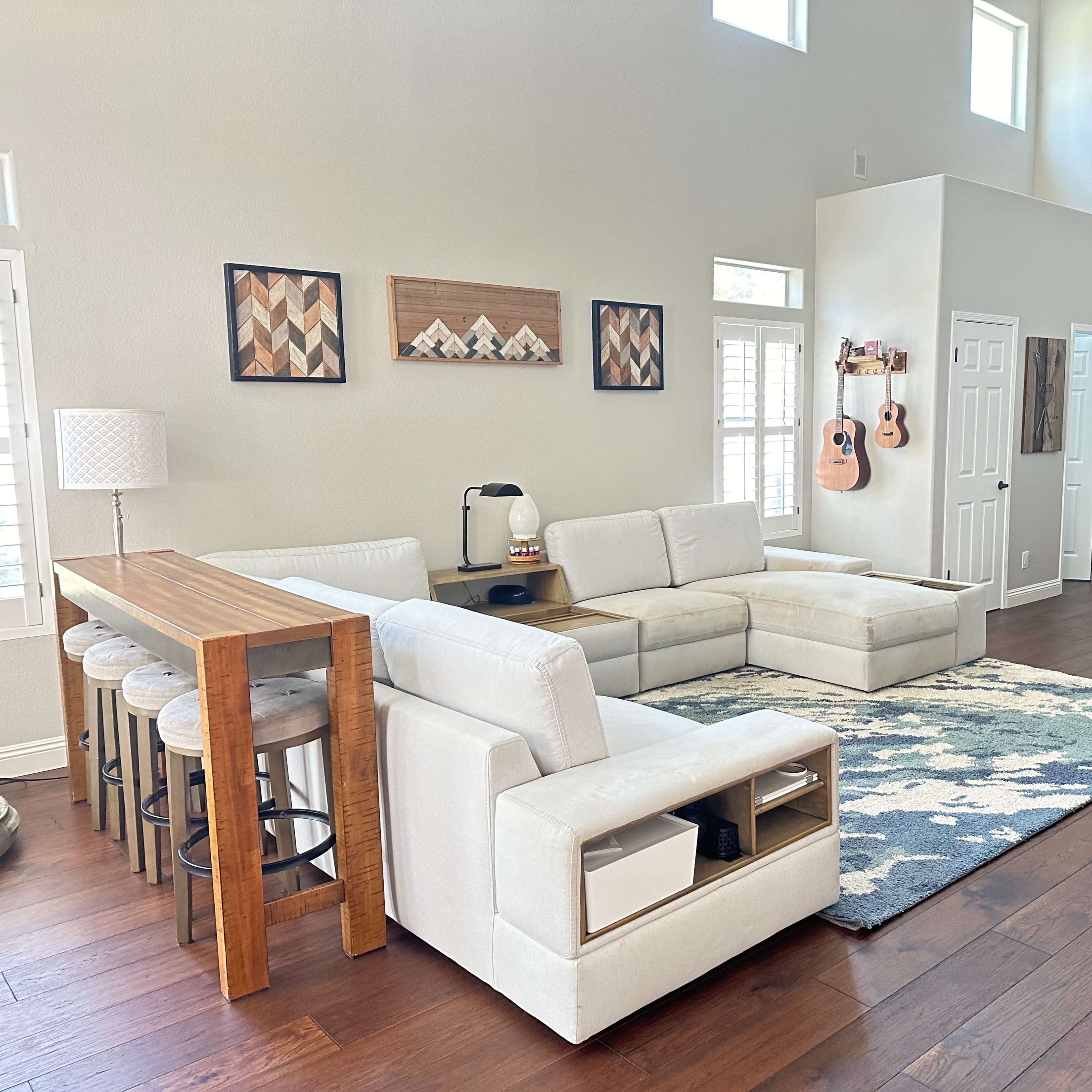 The spacious living room features a sectional sofa, a wooden dining table with stools, a decorative rug, and a staircase leading to an upper level.