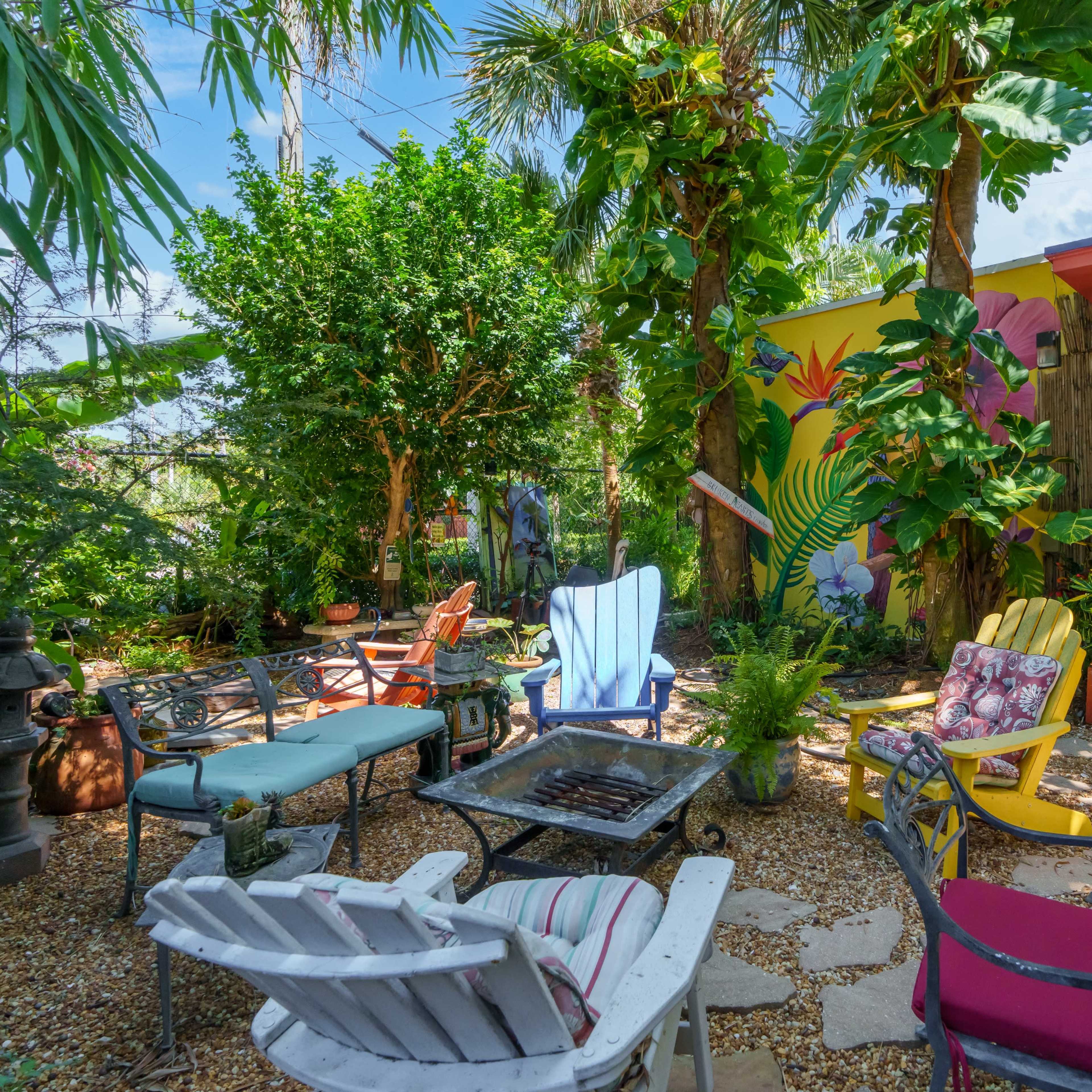 A vibrant garden setting with various colorful chairs arranged around a fire pit, surrounded by lush greenery and a mural on a nearby wall.