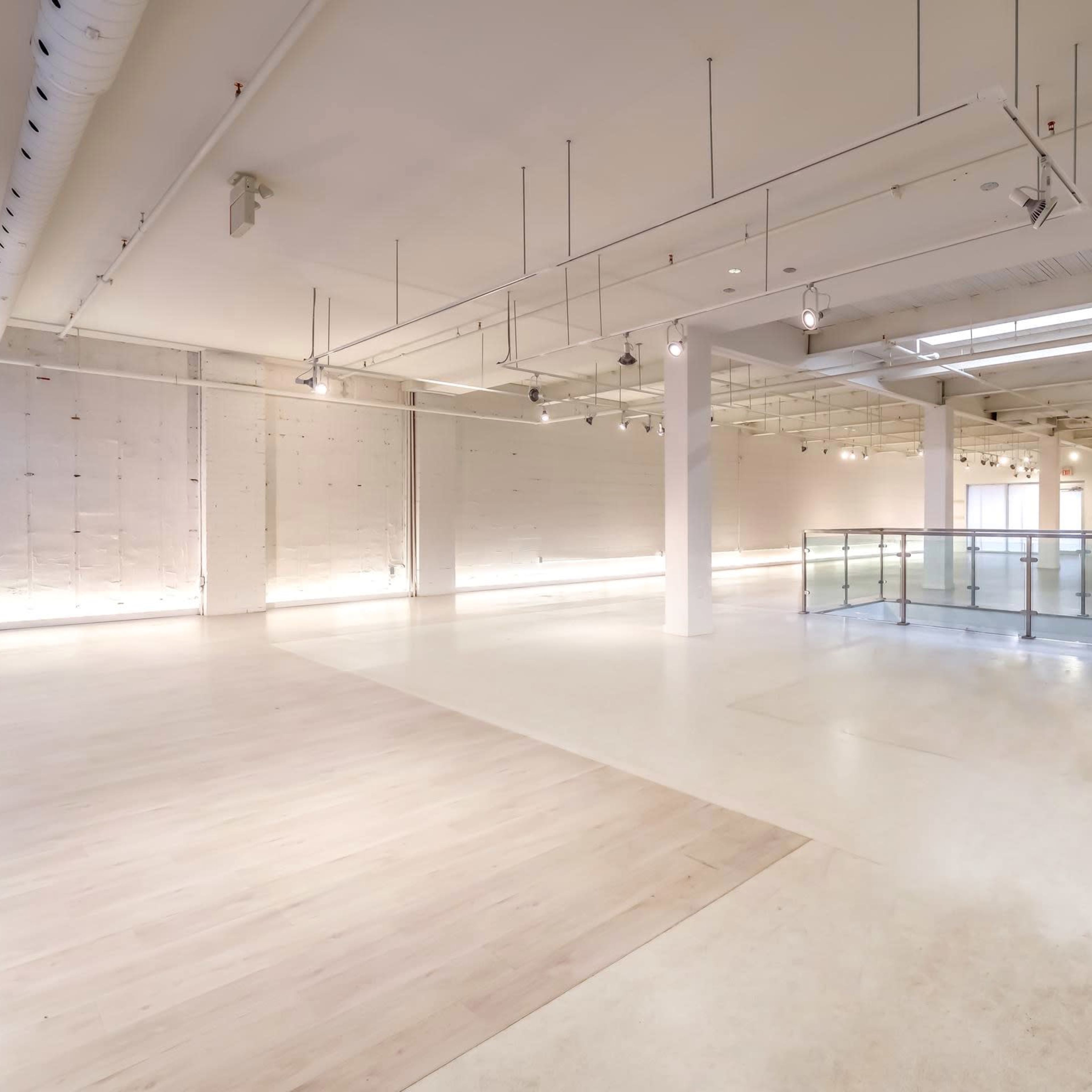 A spacious, well-lit indoor area with white walls, wooden flooring, and a glass railing overlooking a lower level.