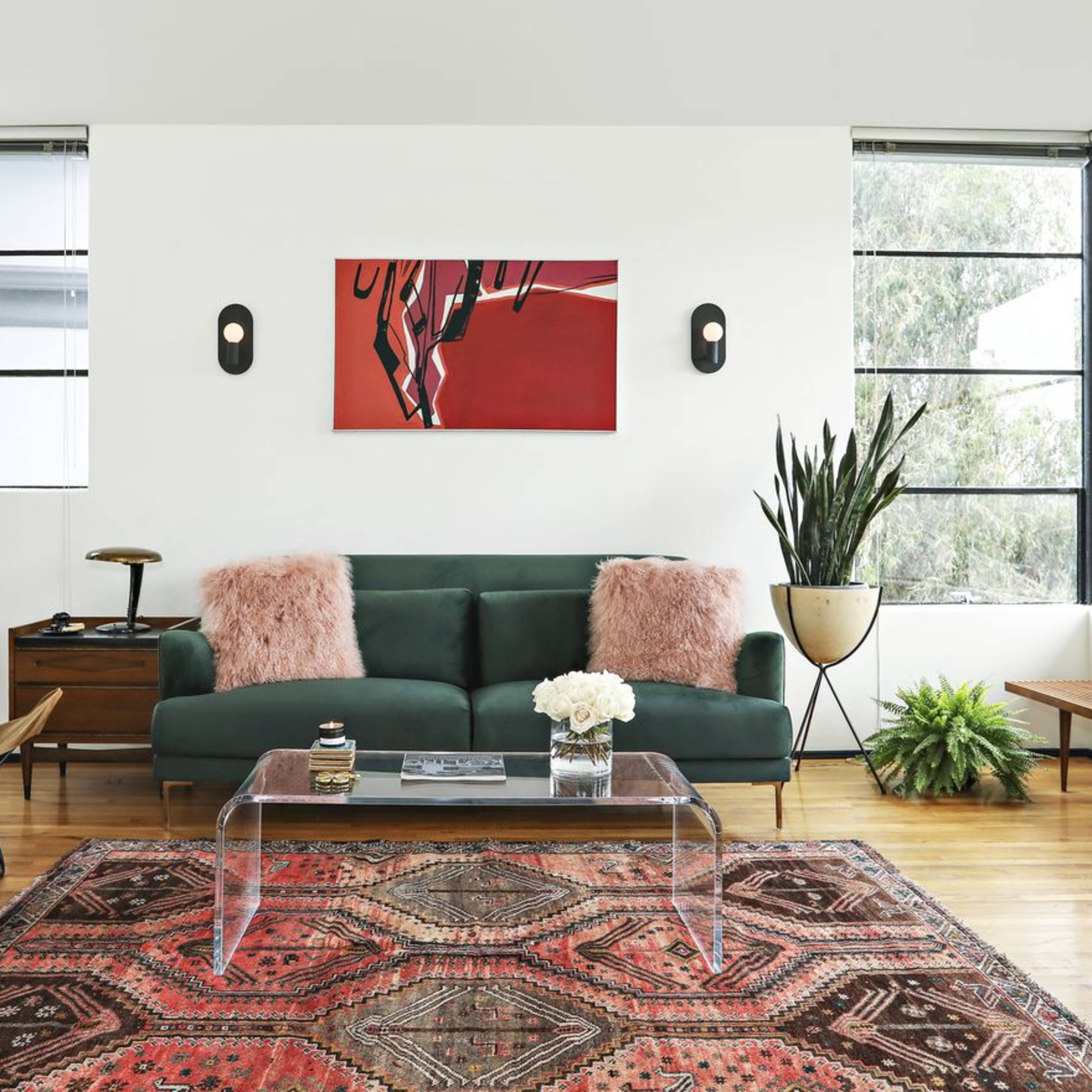A modern living room features a green sofa with pink cushions, a glass coffee table, an abstract painting on the wall, and various plants in the corners.