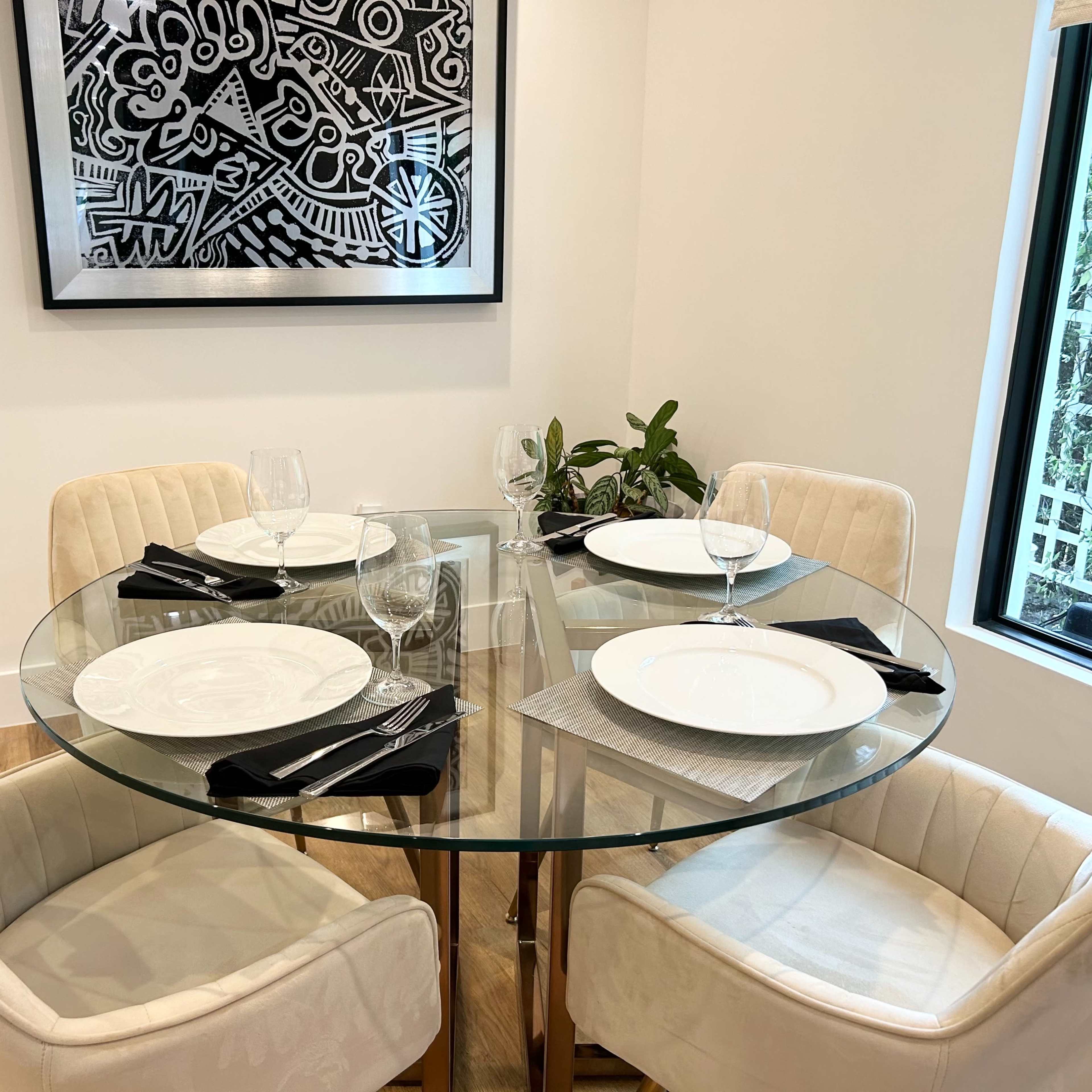 A round glass dining table is set with four plates and wine glasses, surrounded by cream-colored chairs in a modern room featuring a black and white artwork on the wall.