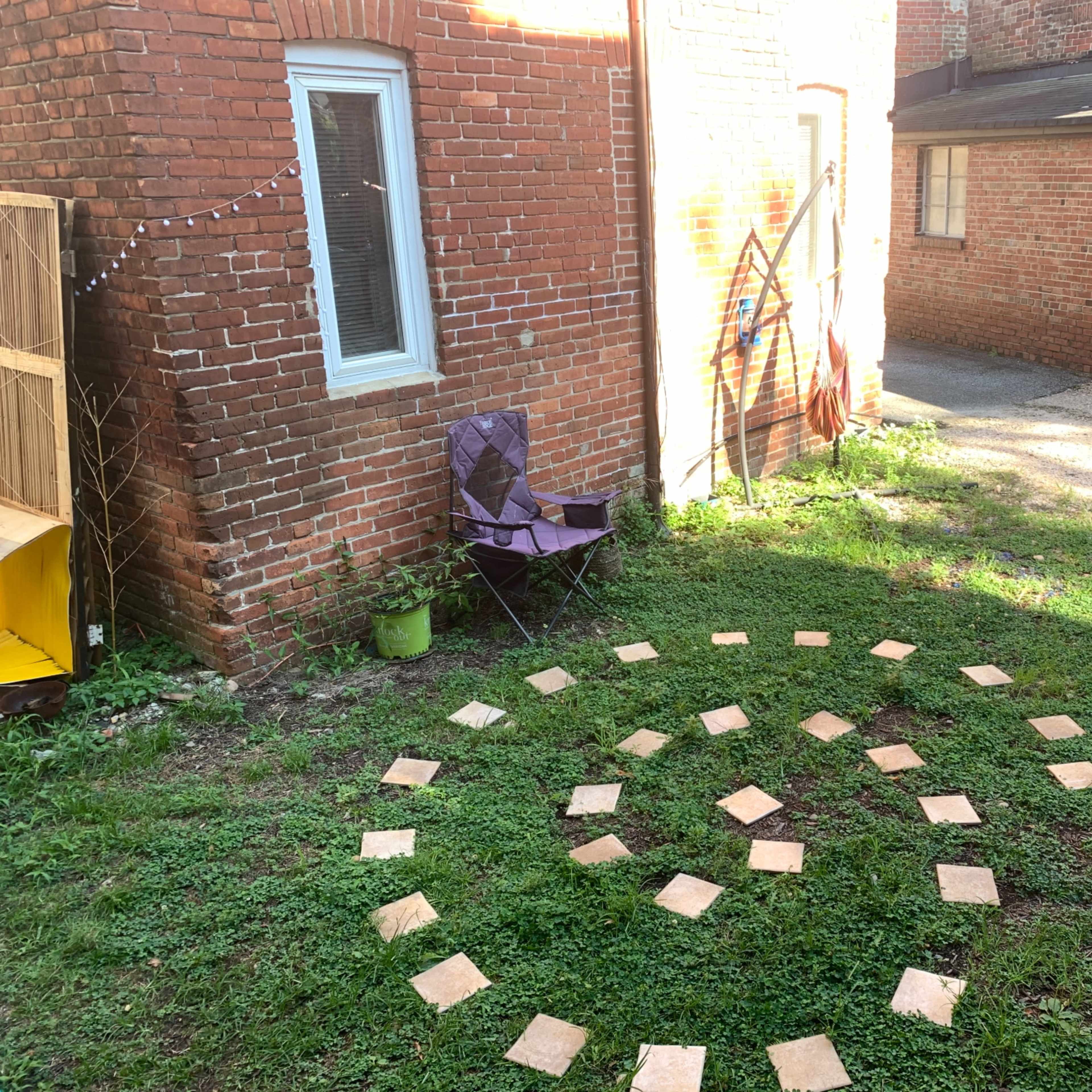 A grassy area with a spiral pattern made of square tiles surrounded by a purple folding chair and red brick buildings in the background.