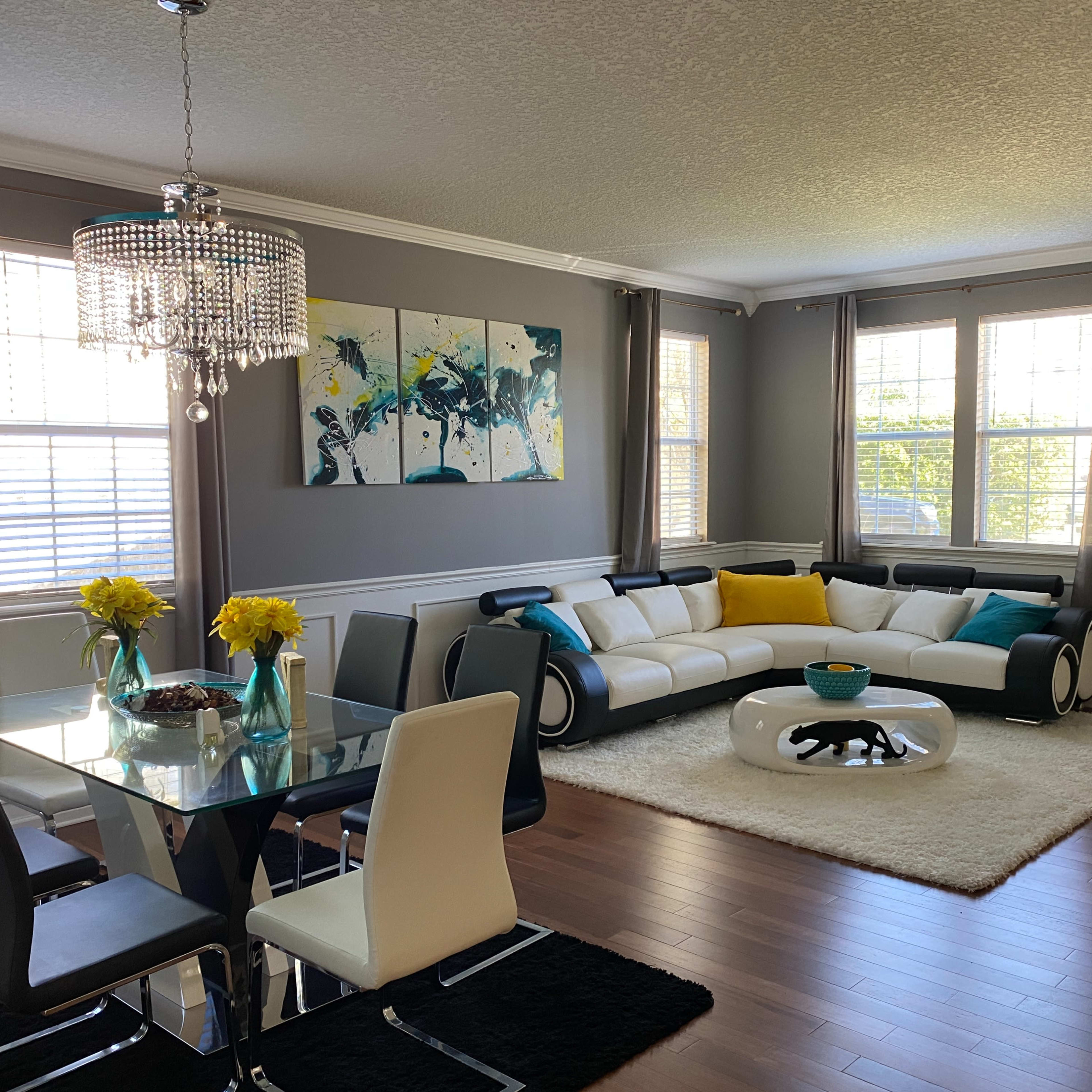 A modern living and dining area with a large sectional sofa, a dining table set for six, and a chandelier overhead, accented by a colorful wall art piece.
